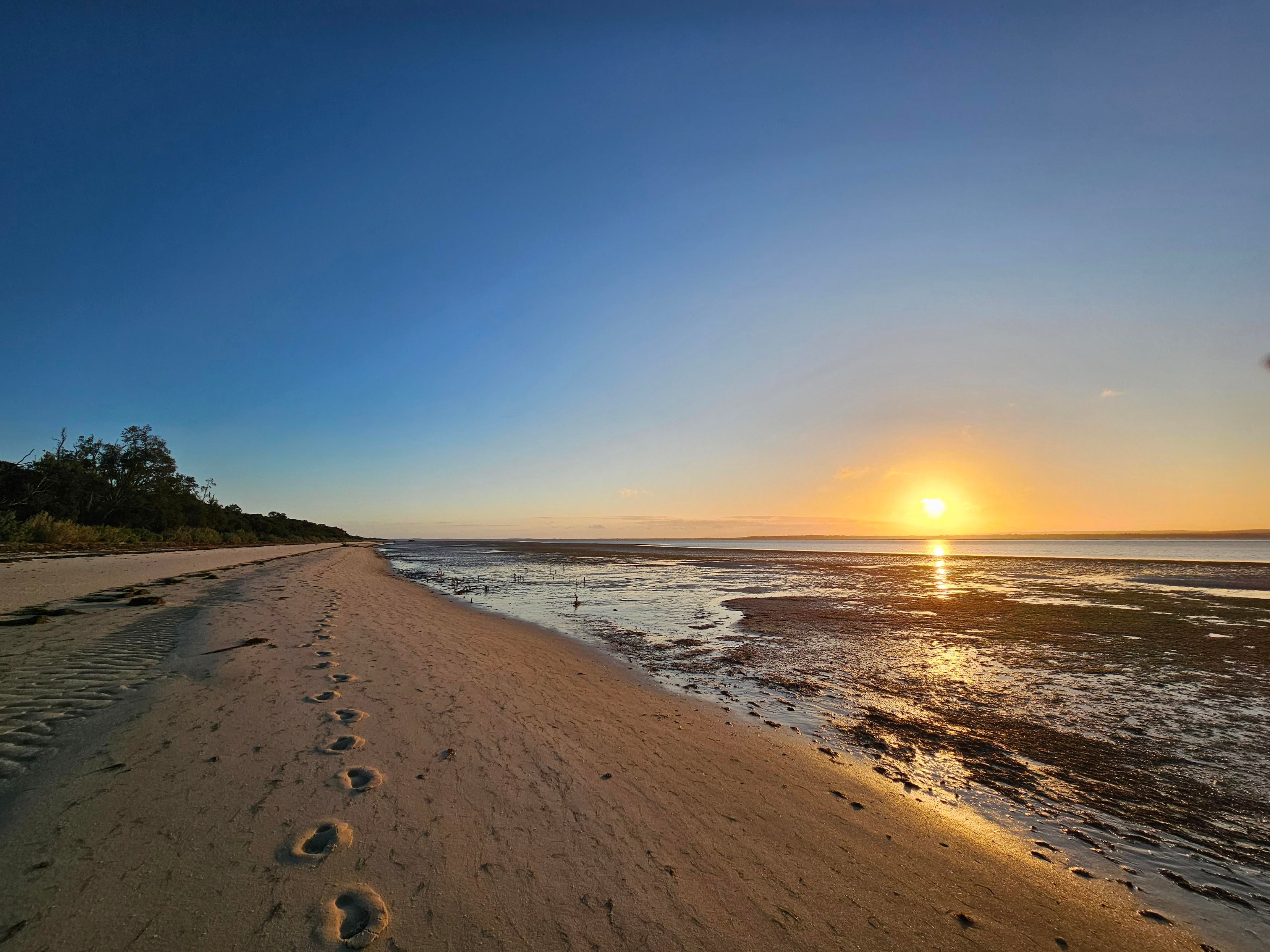 A shot of the (French island) beach at sunset. Clear blue sky, setting sun reflected in the water, golden sands stretching into the distance marked only by my footprints.