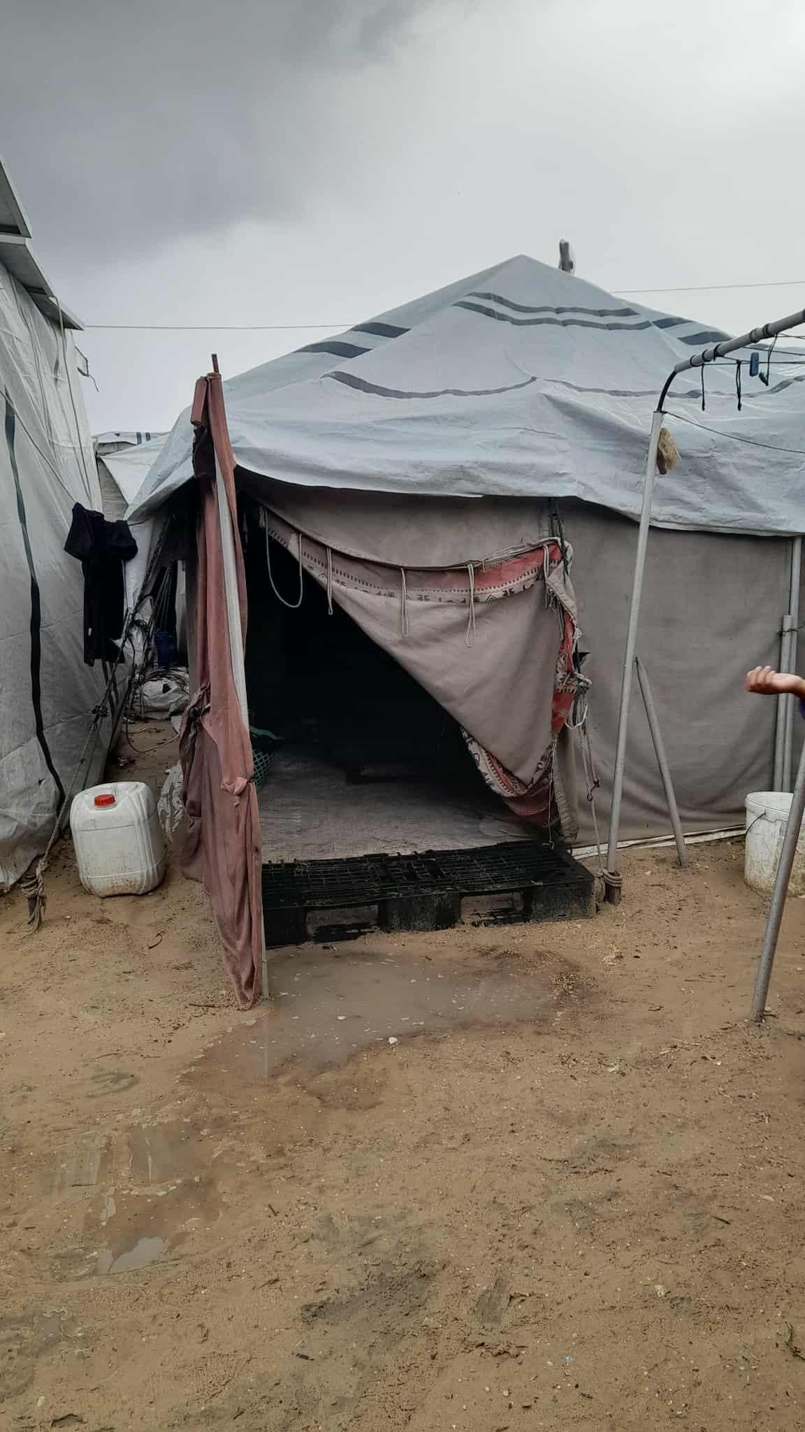 A makeshift shelter, likely a refugee tent, with a partially open flap revealing a dark interior, constructed of grey tarpaulin, positioned on a sandy, muddy ground, with other similar tents visible in the background.