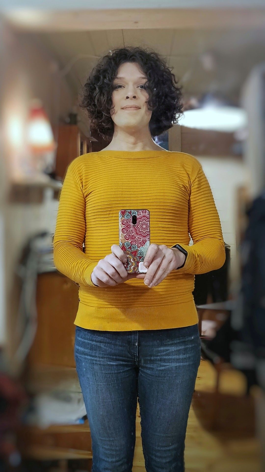 Mirror selfie taken from below the chest, wearing a yellow shirt and blue jeans, head held high, smiling and looking down into the camera