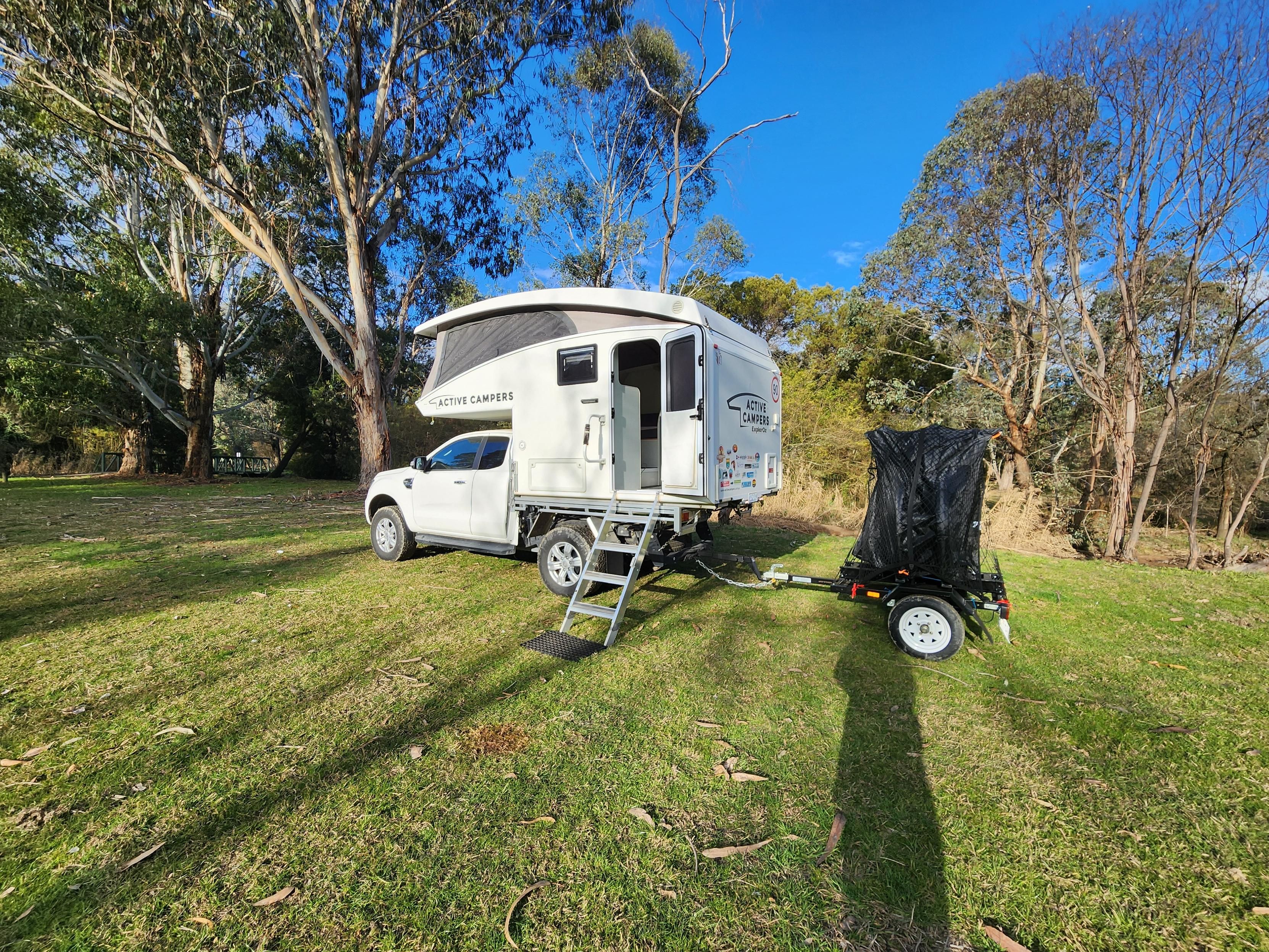 Slide on camper (Rhonda) with top up, atop a white ute (Ronnie), in late afternoon winter sun. There is a dinky bike trailer behind, with 2 wrapped e-bikes. 