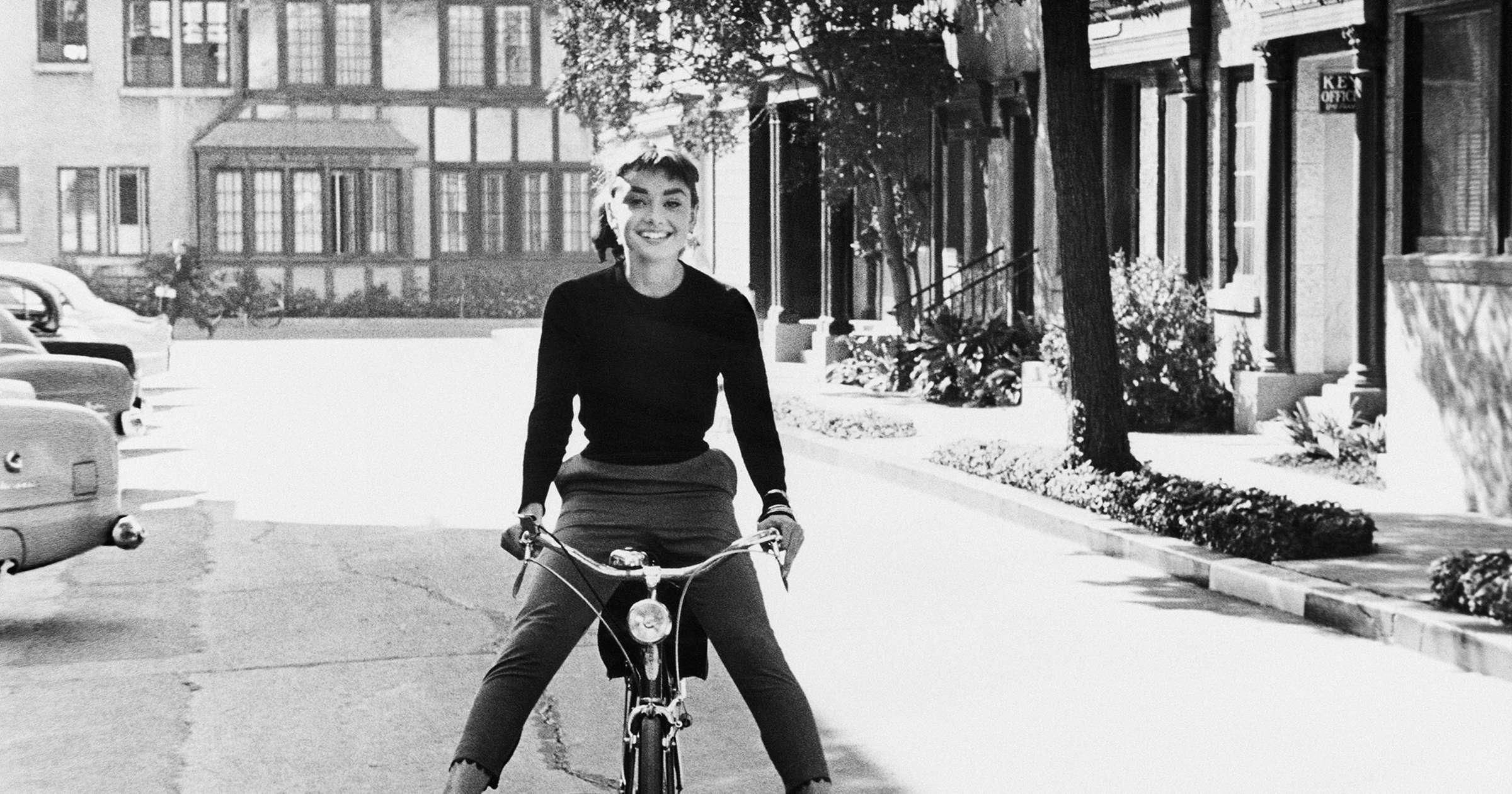 Audrey wears a black top and dark pants, and is seen head-on as she sits on a bicycle, smiling. 