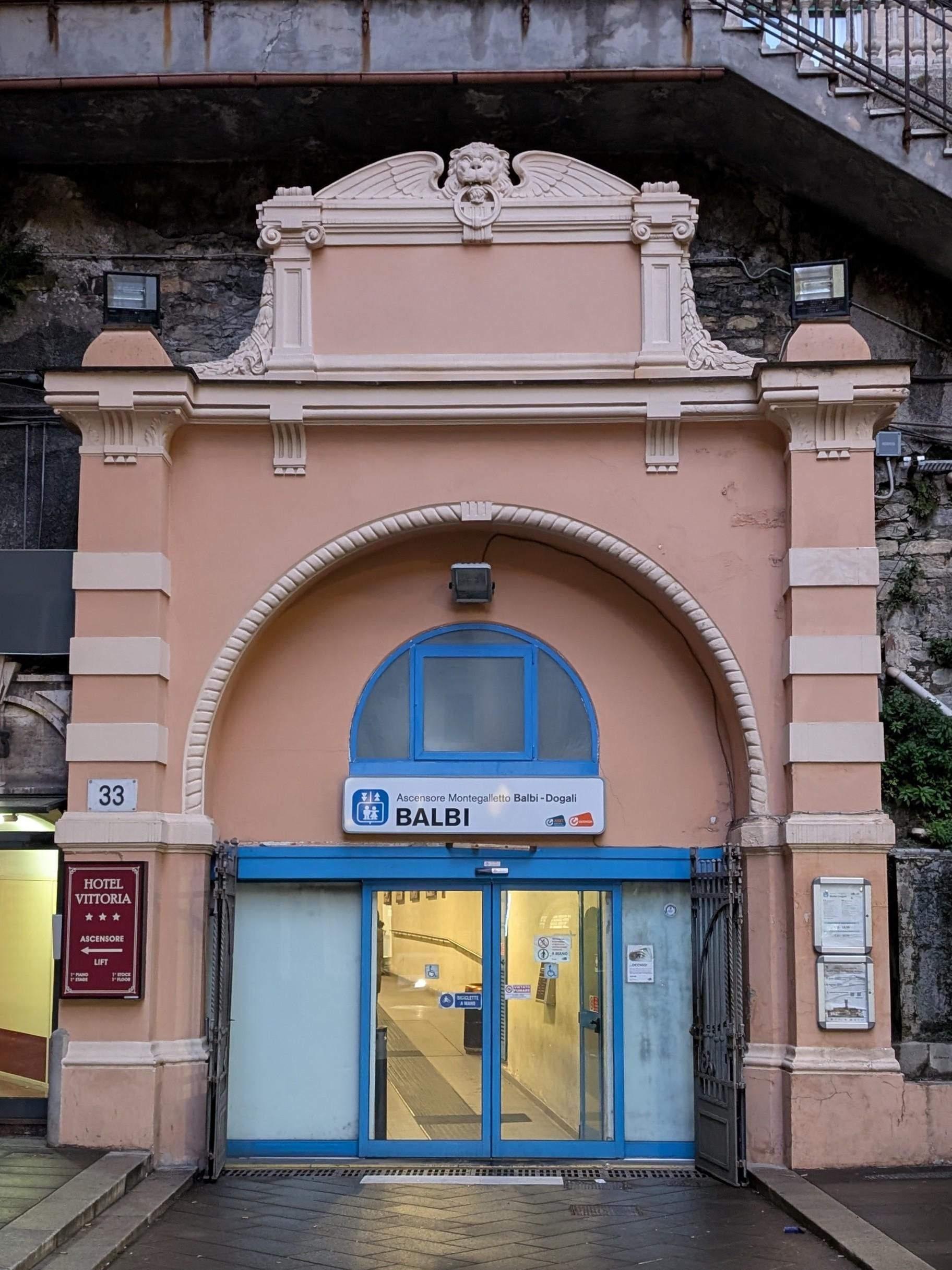 The arched entrance with salmon-pink façade with ornaments, glass door with blue doorframe, rock face, overhead sign "Ascensore Montegalletto Balbo-Dogali"