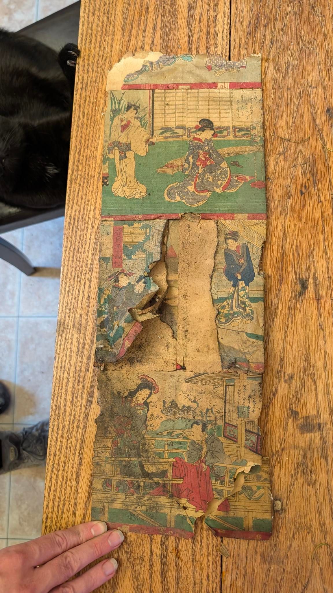 A very yellow and eaten parchment sits on a wooden table. You can see traditional Japanese style images and some very small text. The middle is torn, missing and the whole thing looks very fragile.

A hand is holding down it's curling lower edge.