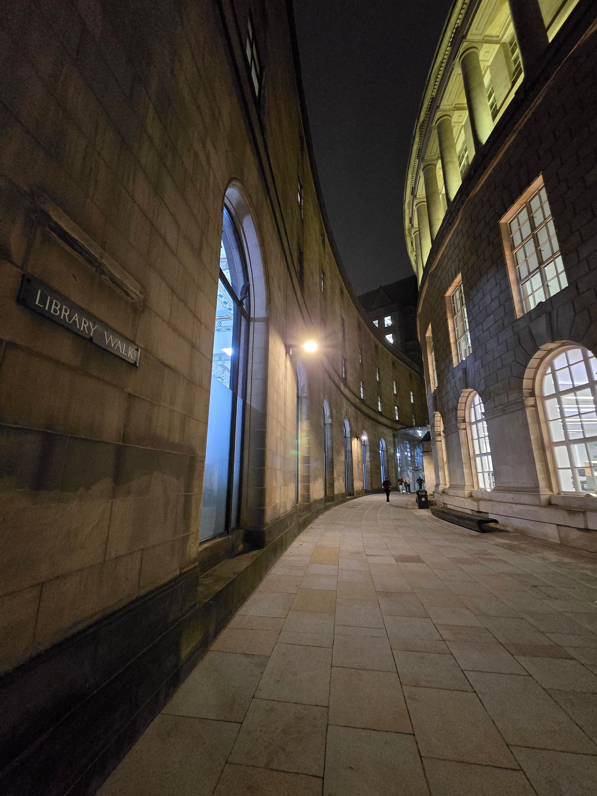 night photo of a curved alley between buildings in Manchester with a sign on the wall with text "library walk"