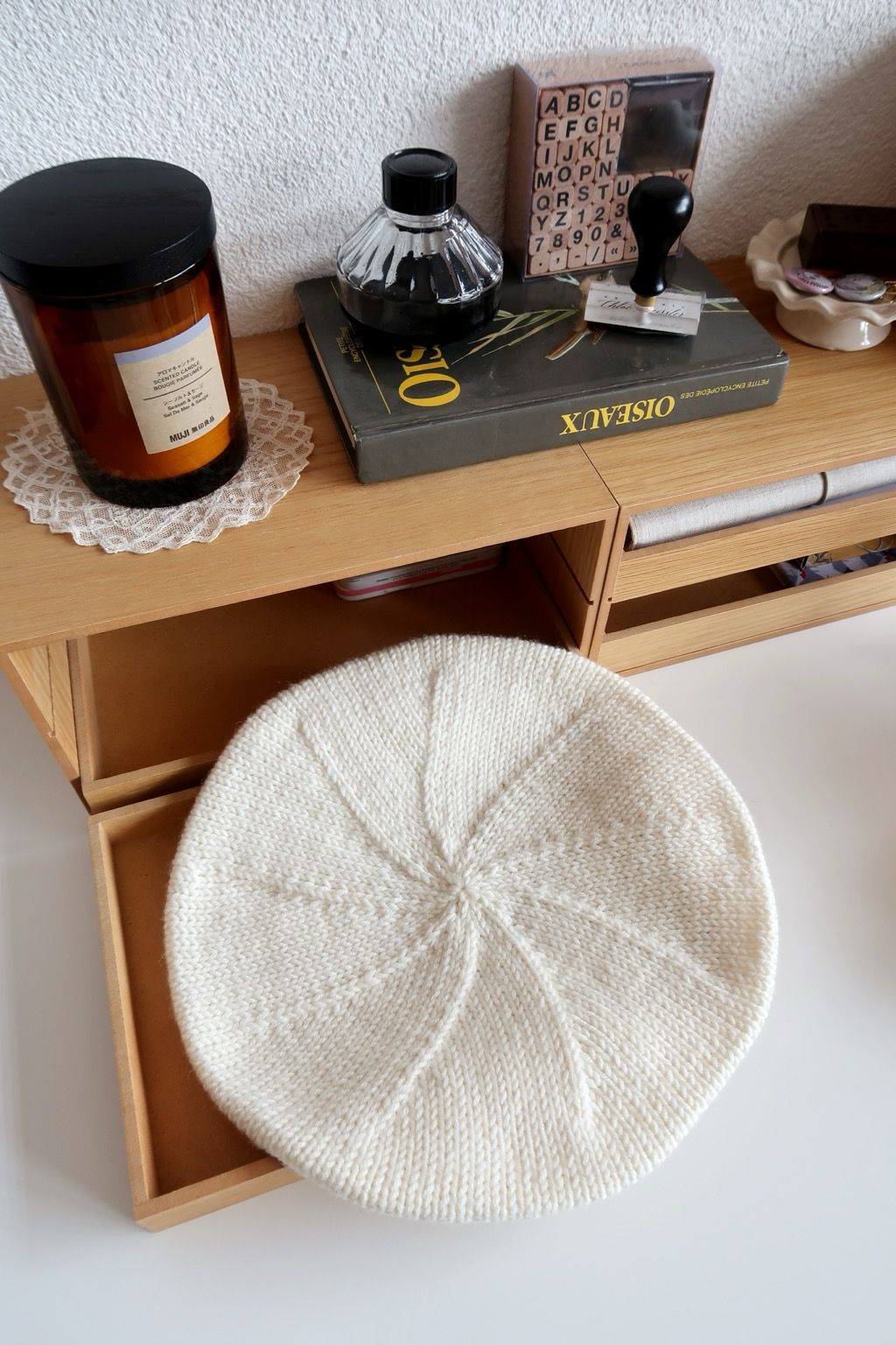 Un béret en tricot vu d’en haut, posé sur une petite étagère en bois dont le tiroir est ouvert. Autour, dans le décor, il y a une bougie posée sur un napperon en dentelle, un livre d’ornithologie ainsi qu’un pot d’encre noir, un tampon encreur et un kit alphabétique de mini tampons encreurs.