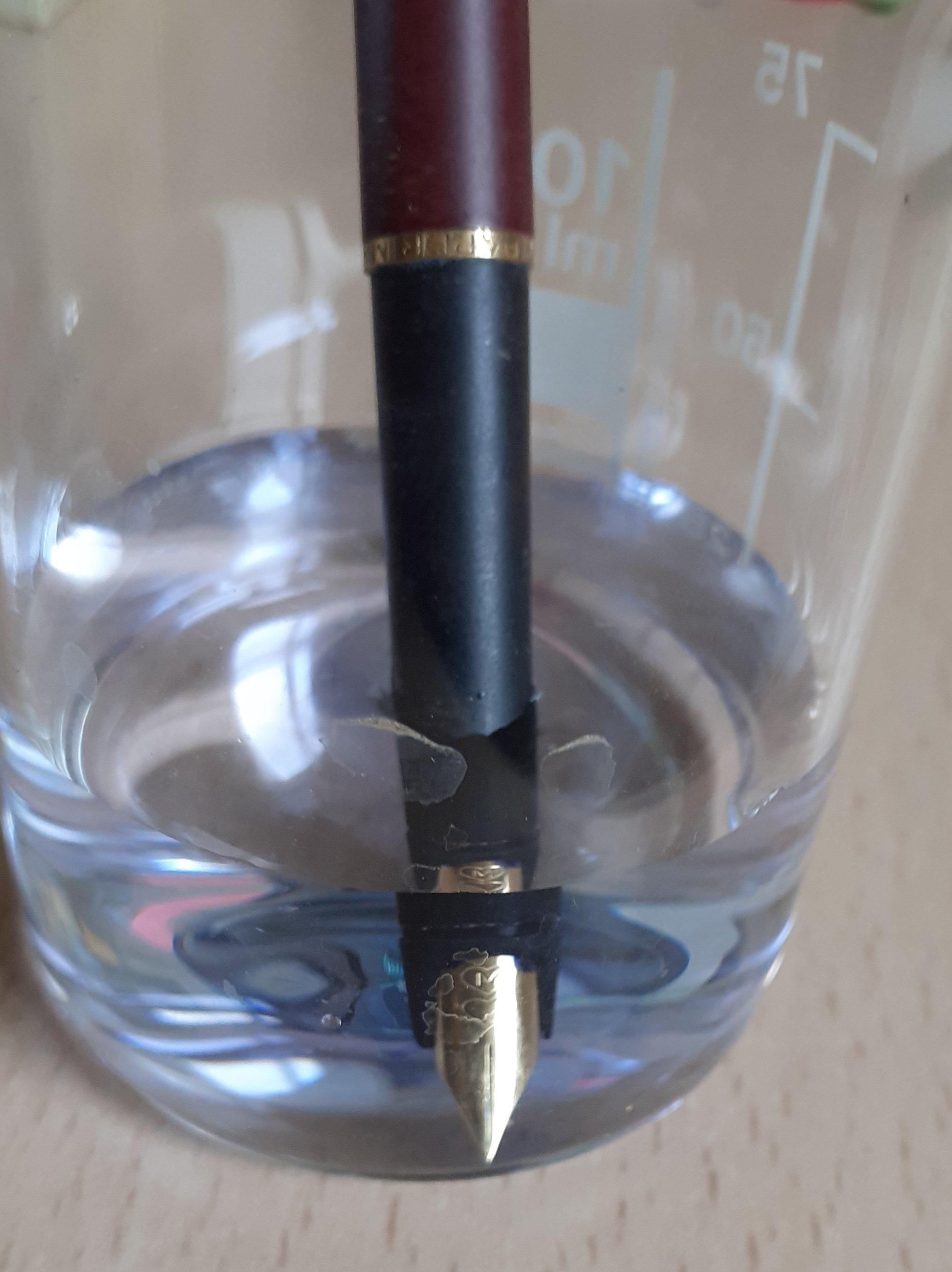 La foto muestra la punta de una pluma estilográfica sumergida en un vaso con un poco de agua. El agua tiene una tonalidad ligeramente azul debido a la tinta que se ha disuelto. El plumín es dorado y tiene grabado el lodo de la marca, dos corazones uno sobre el otro. La zona de agarre es de color negro y el cuerpo de la pluma es granate, separado de la zona de agarre por un disco dorado.