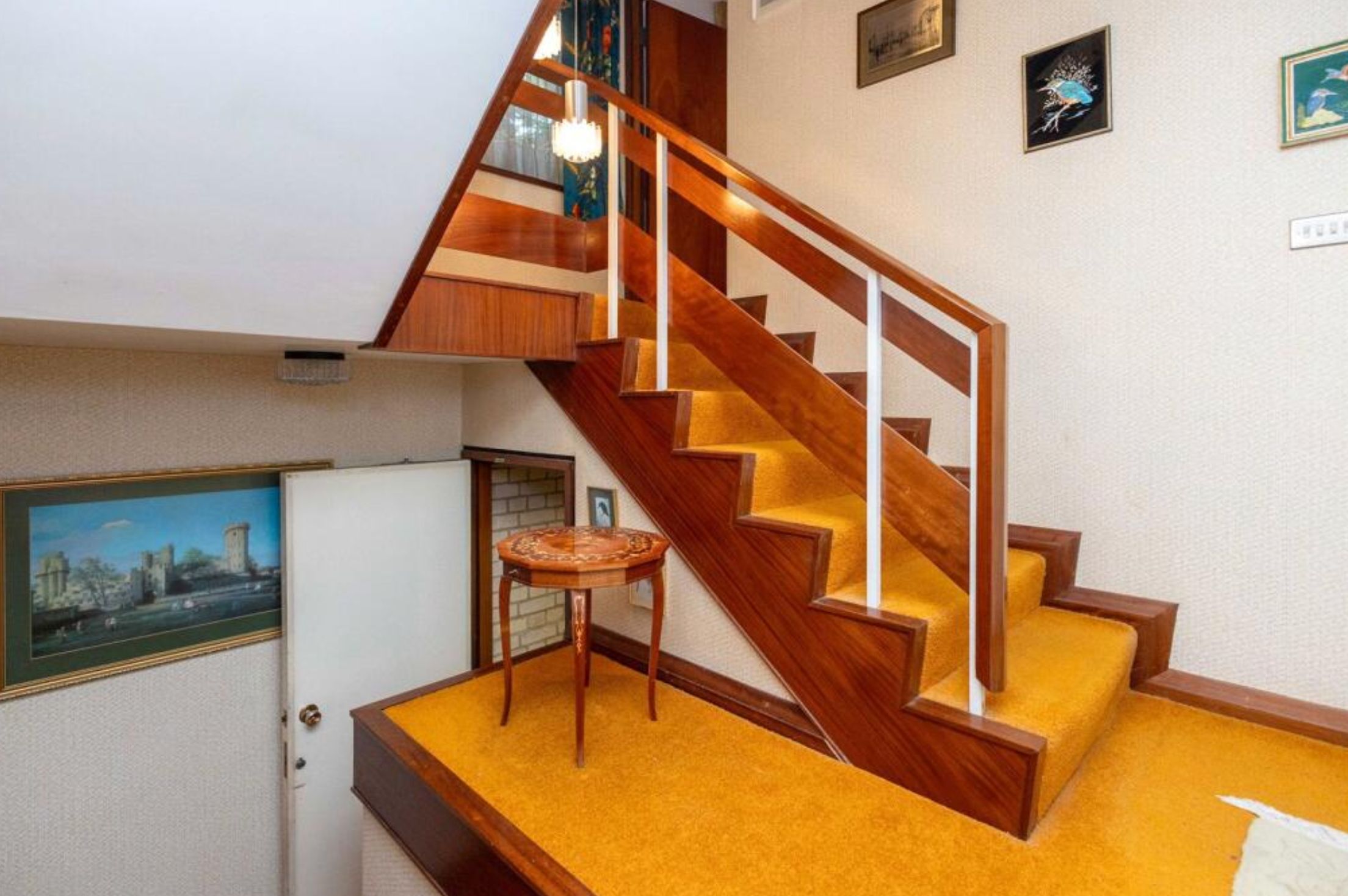The landing of some stairs with orange carpet. Alongside the stairs is a small landing with no railing, and a side table positioned towards the back of it. 