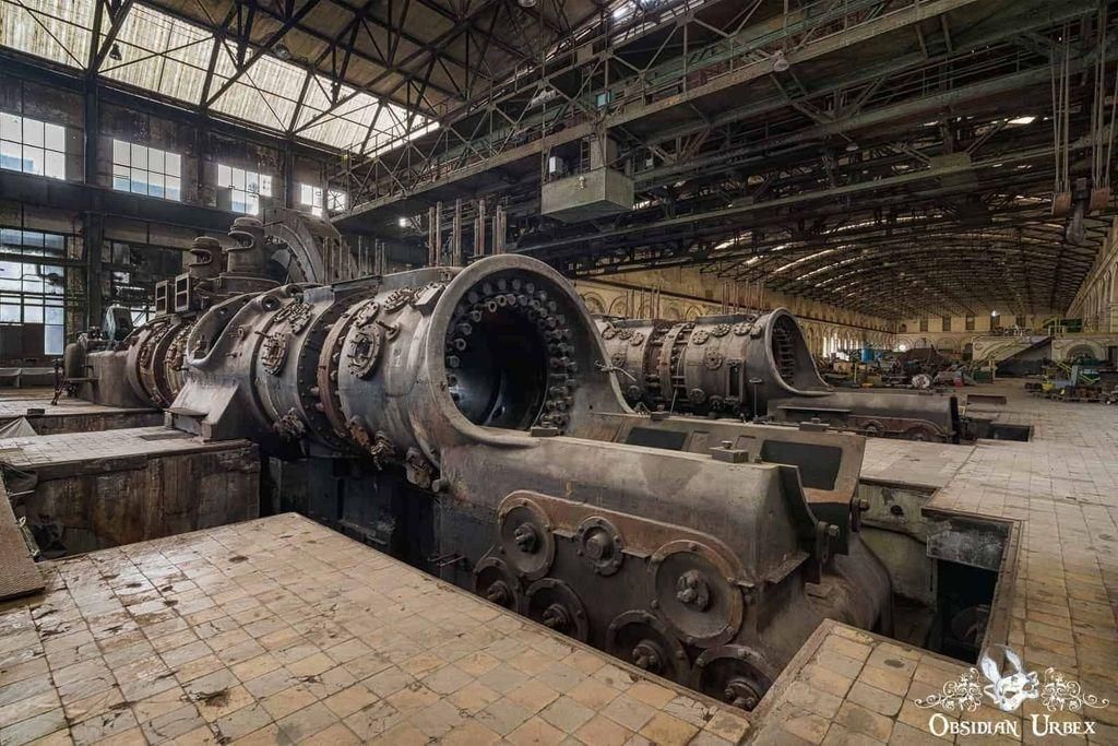 Inside a factory-like building, two enormous and ornate rusting machines rest on tiled floors. Overhead, the metal beams of the large building are visible