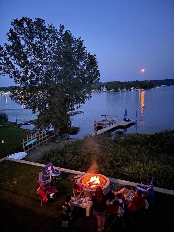A photo of a lake, a boat ramp, some people sitting in the foreground and a big beautiful moon!  The moon is reflecting on the water in an orange line.