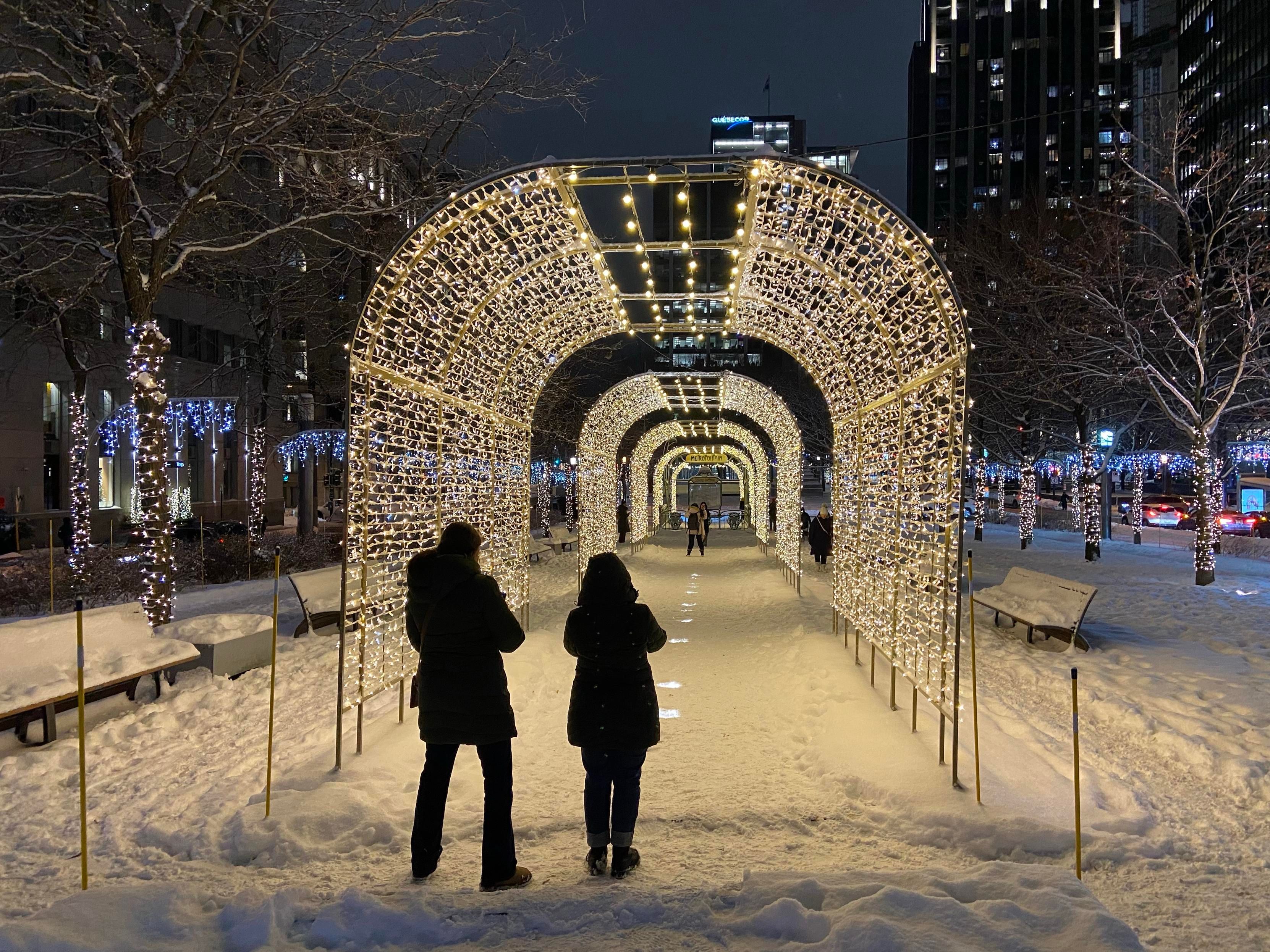 Nighttime in montreal, two people are standing taking photos in front of a long snowy corridor covered by an arch of fairy lights. Further along, under the arch, are two more people taking photos. 