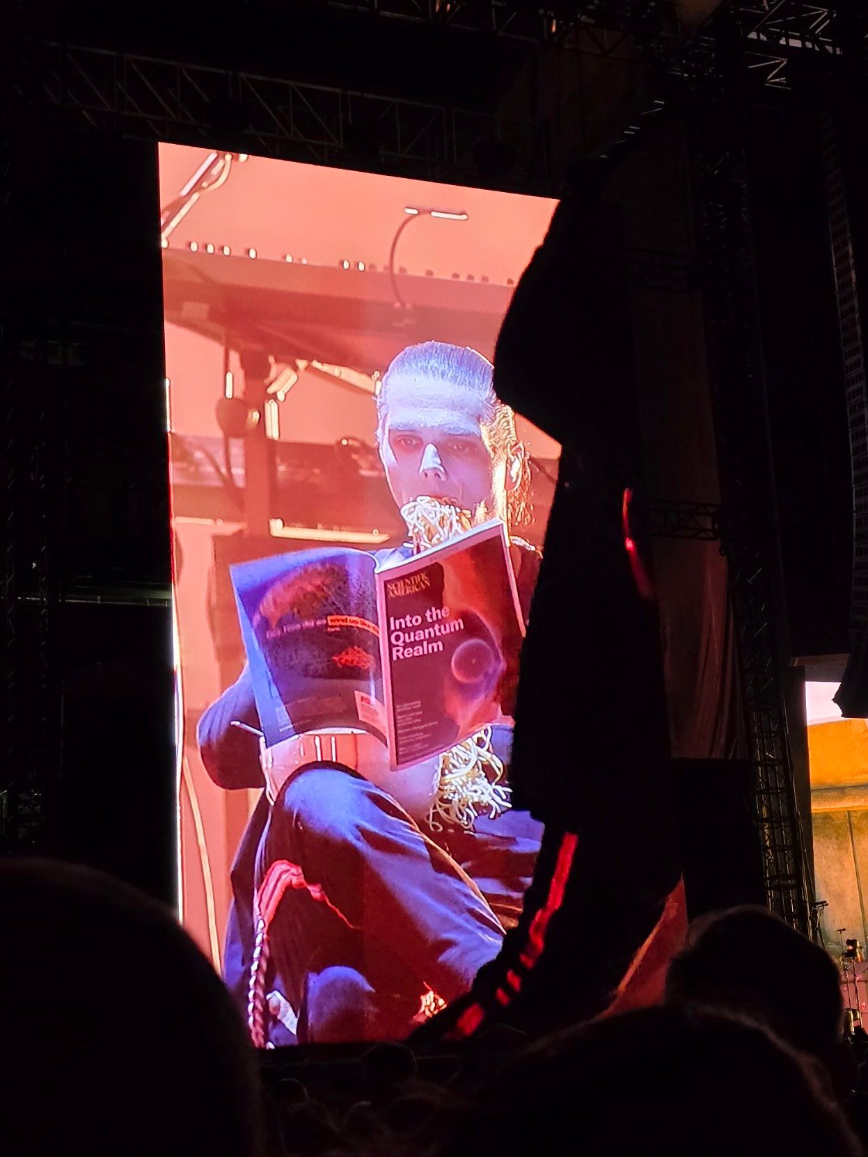 Gerard Way eating spaghetti while reading a magazine on stage.