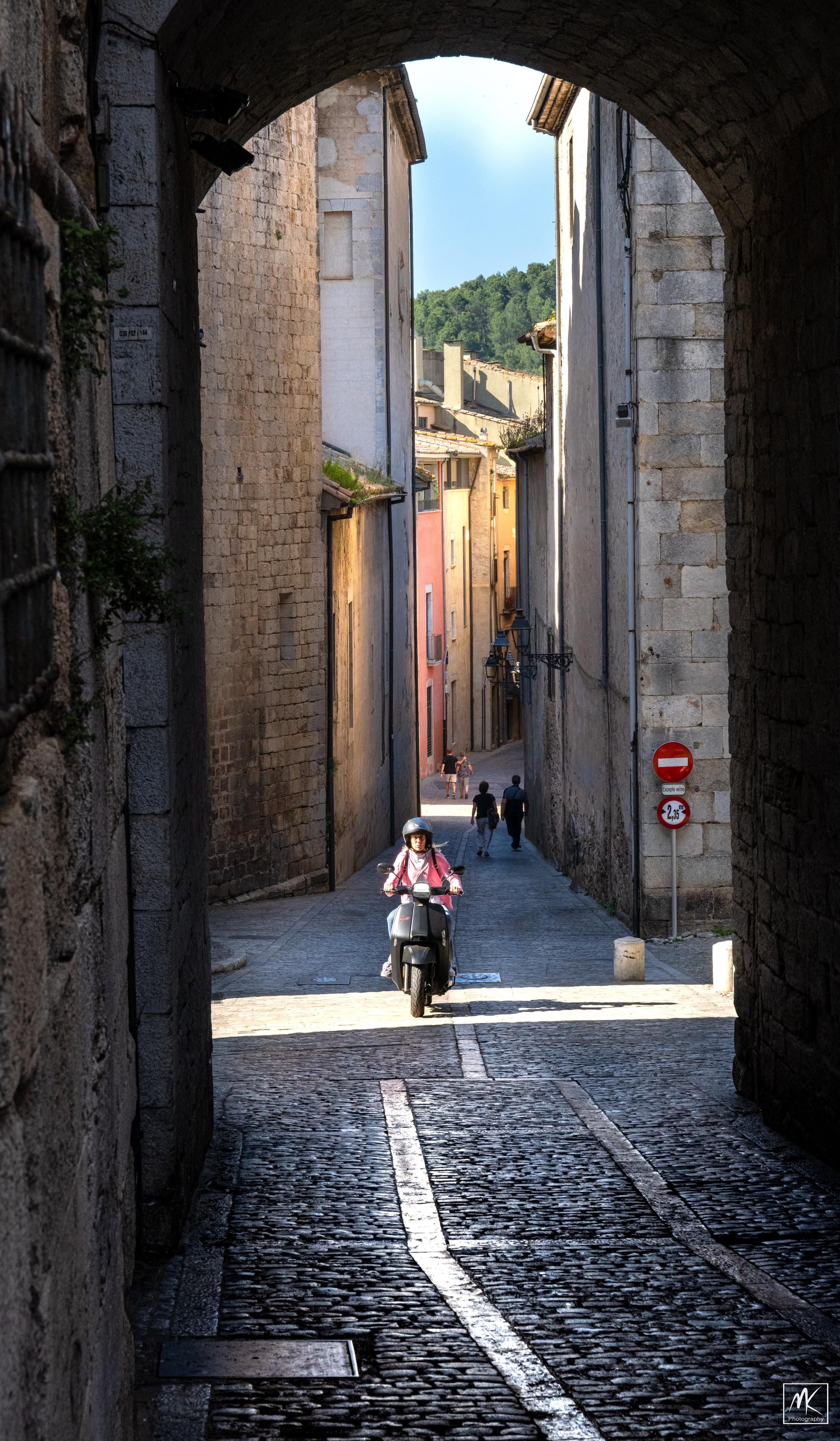 Color photo looking down a cobblestone street passing through a dark archway, with a person on a motorcycle in the sunlight beyond and the street curving away between buildings behind them. 