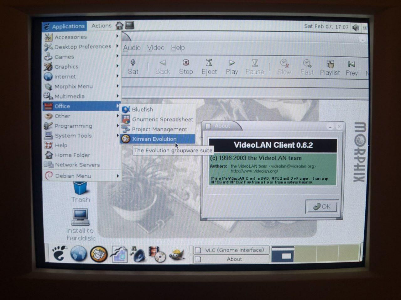 A photo of a CRT screen. Gnome menu shows that there are quite a few programs available. There's Video LAN 0.6.2 running in the background, with horrific fonts that are barely readable
