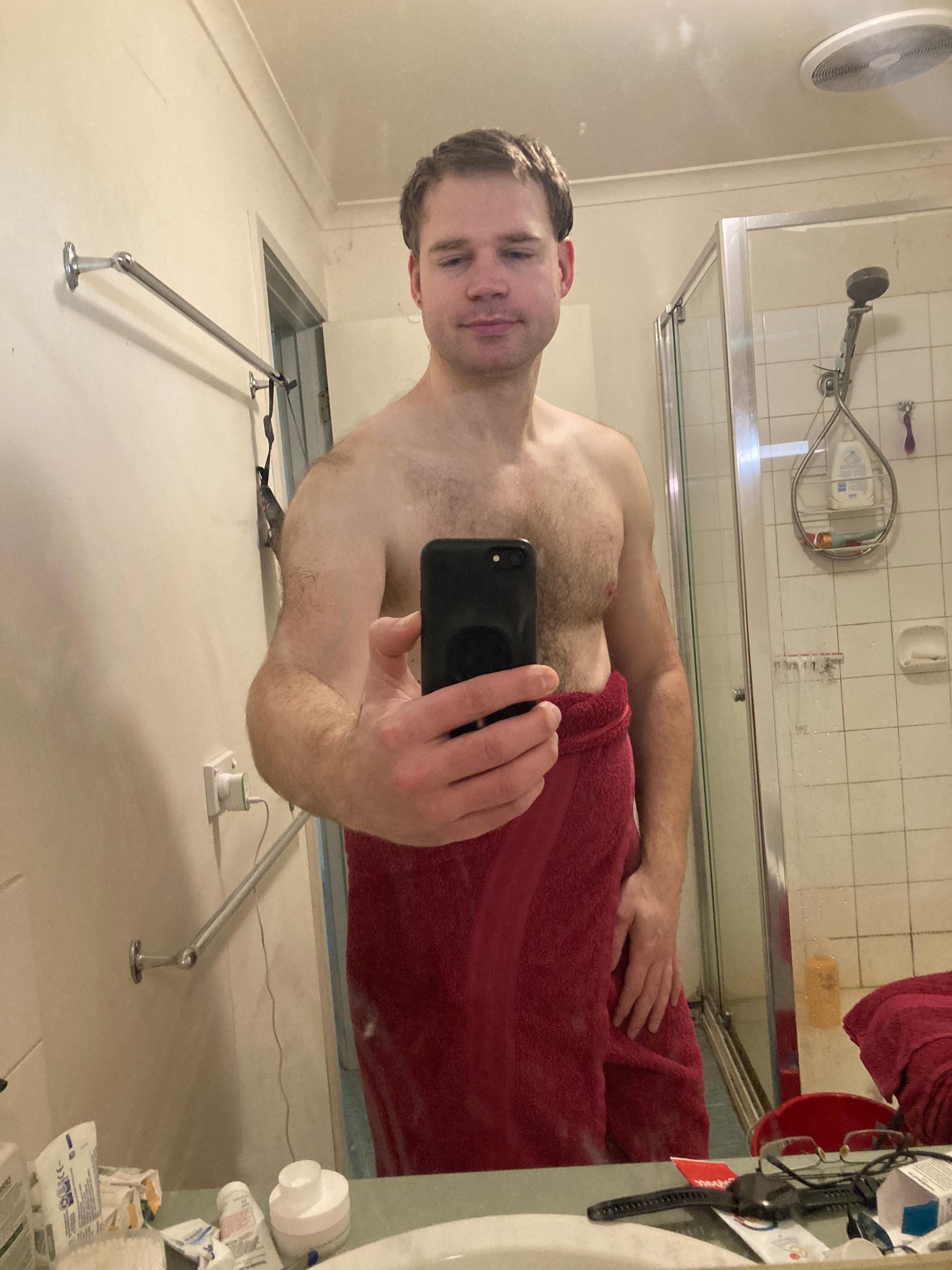 post-shower bathroom selfie of Luke. They are smiling at the camera and showing off their chest. They are wearing a red bath towel around their waist.