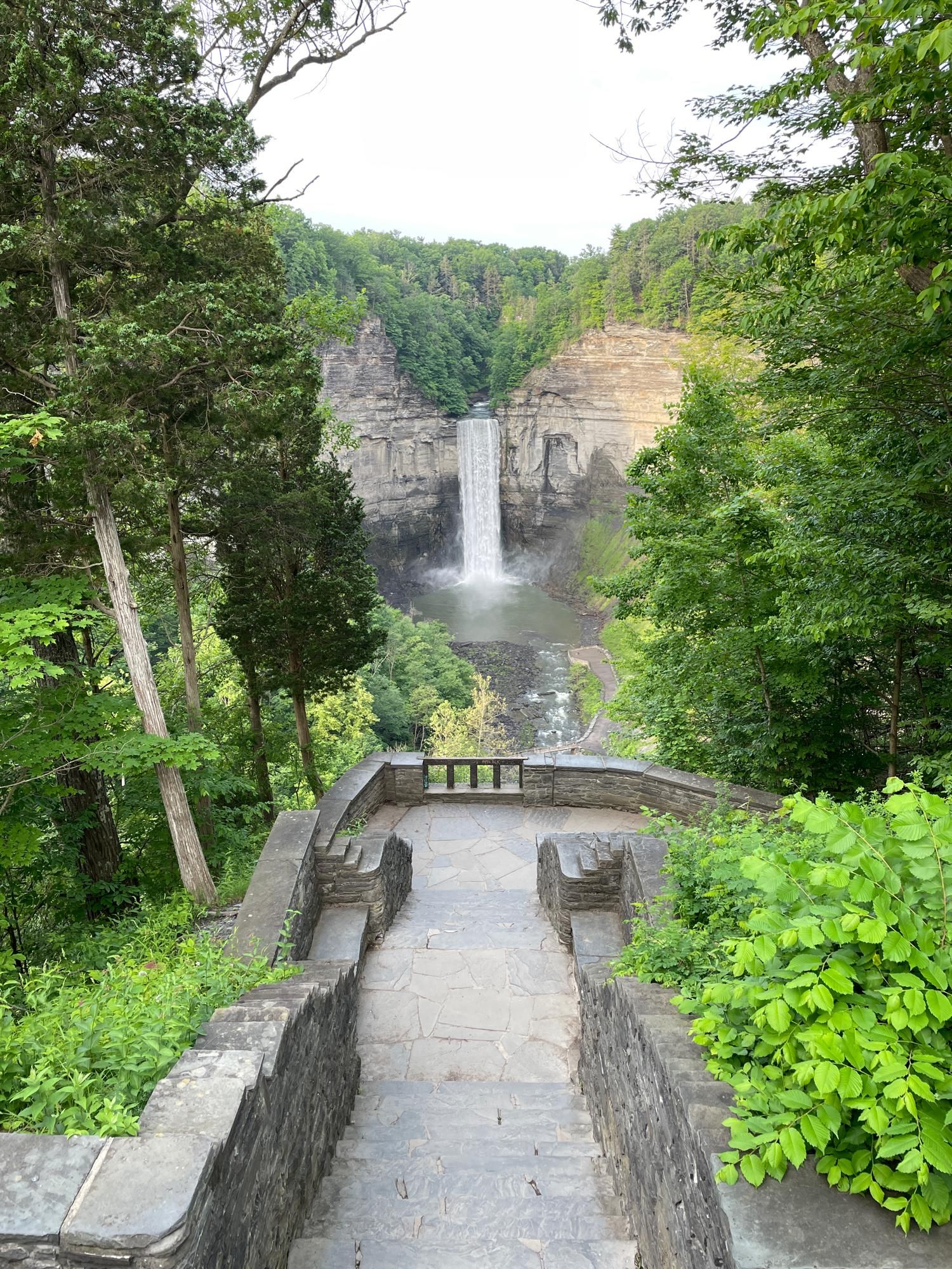 Beyond a stonewalled overlook is a tall waterfall pouring down the rock wall of a gorge.