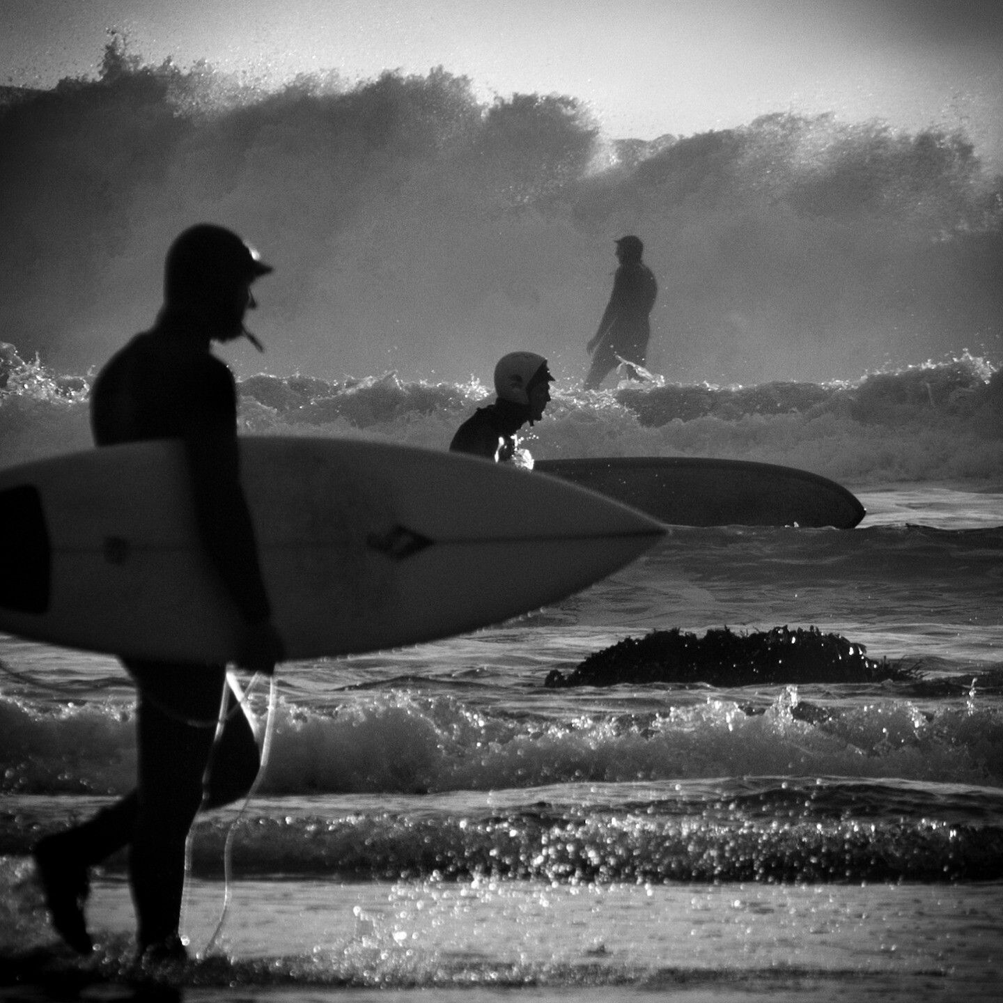 Three levels of surfers: a man in a helmet carrying a board on the shore. Behind him is a man wading into the surf. Behind them is someone riding a wave silhouetted against the large breakers behind them.