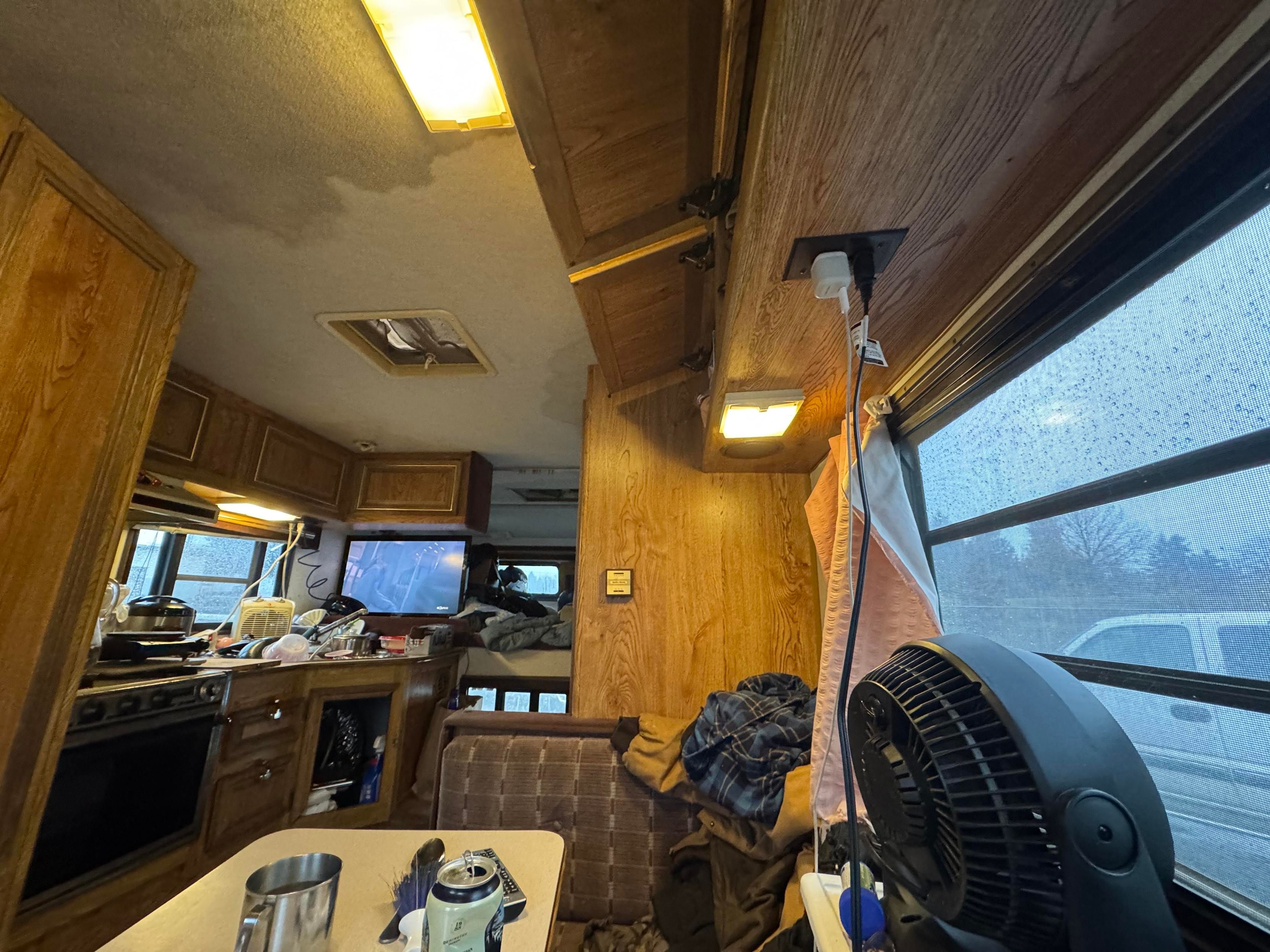 Interior of a camper van showing a small kitchen area with an oven, sink, and television. There are scattered dishes and a fan on a table. The space is lit with a couple of overhead lights, and rain is visible on the window. 