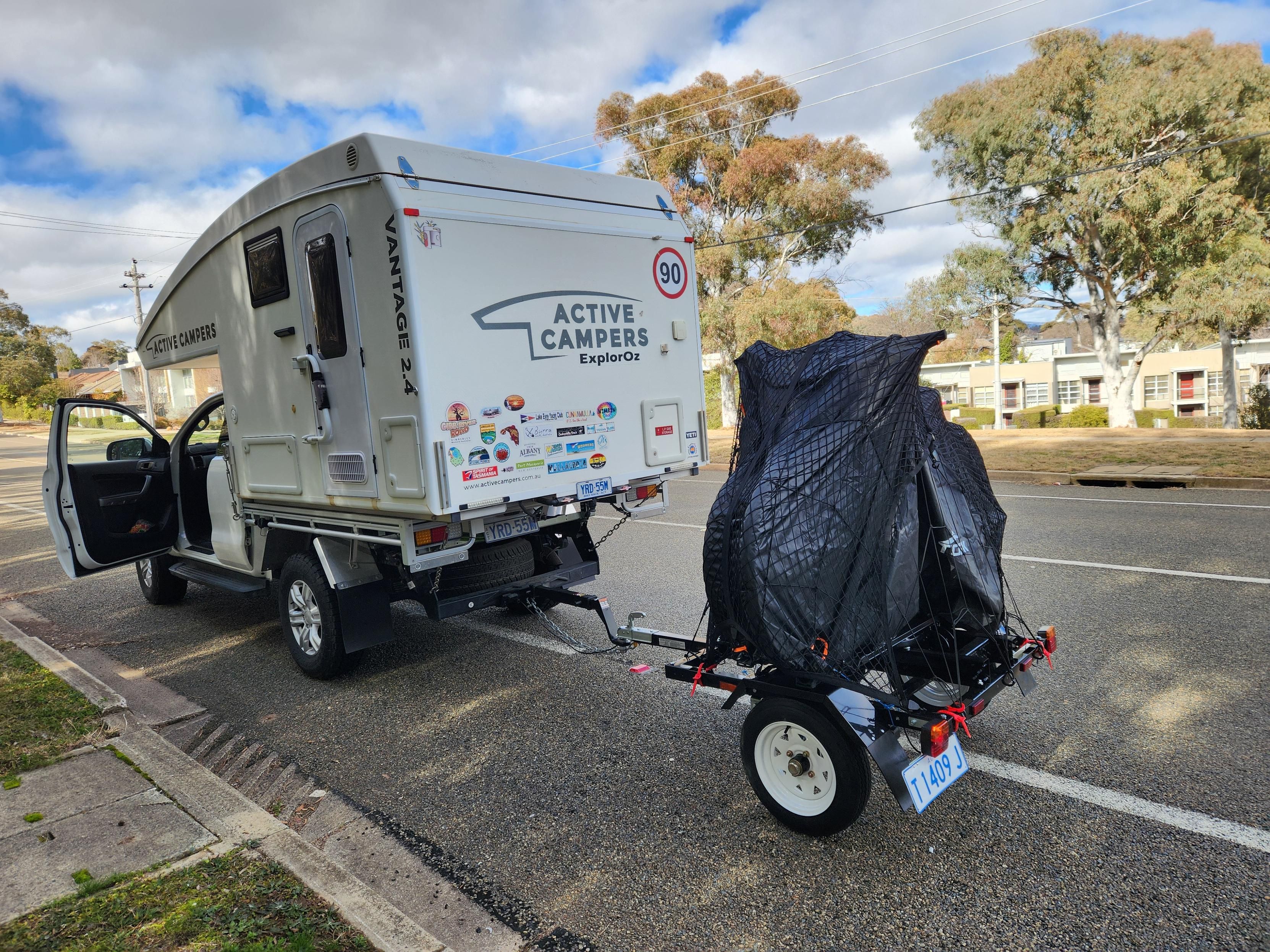 A small slide-on camper atop a white ute with a dinky little bike trailer behind, on which 2 bikes are covered in black wrapping.