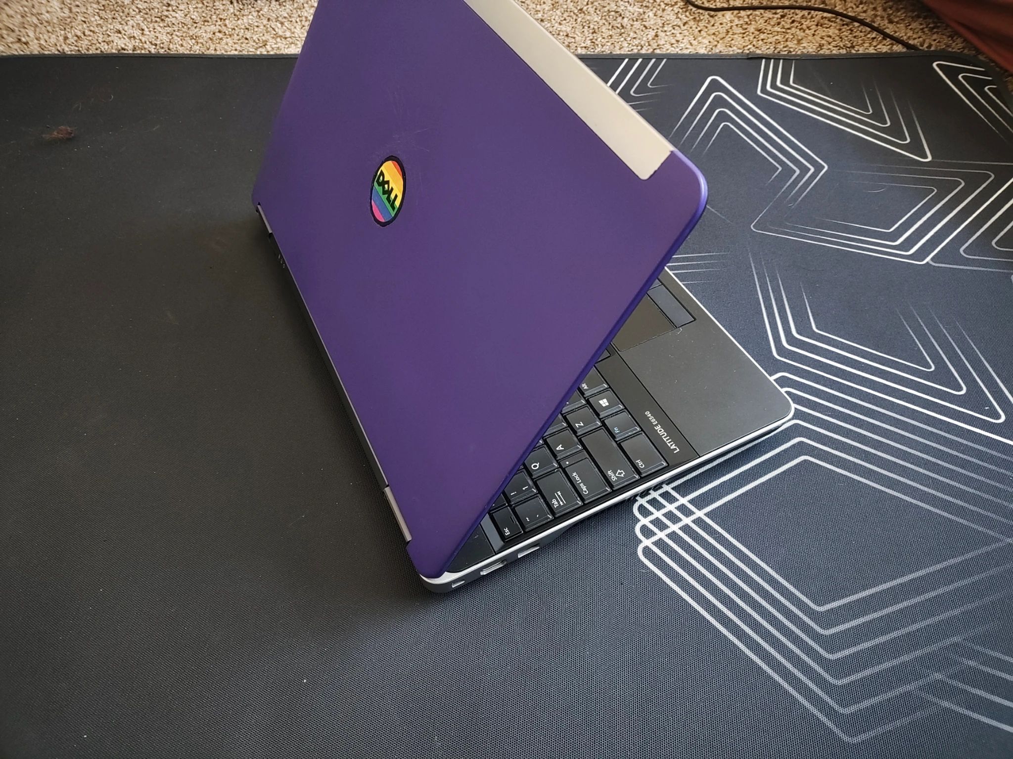 side top back view of a partially open laptop. the inside palm rest and keyboard are black, but the outside shell part is matte purple with a logo circle that says "doll" instead of "dell" and has a horizontal rainbow flag beneath it. 