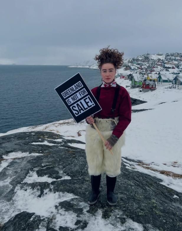 Mia H. Chemnitz stands on a snow-dusted rocky shoreline in Nuuk, Greenland, holding a protest sign that reads “GREENLAND IS NOT FOR SALE!” She wears a dark red sweater, patterned neck scarf, black boots, and traditional light fur pants made from polar bear. Behind her, the grey Arctic sea stretches to the left while a hillside of colorful houses and buildings sits under an overcast winter sky.