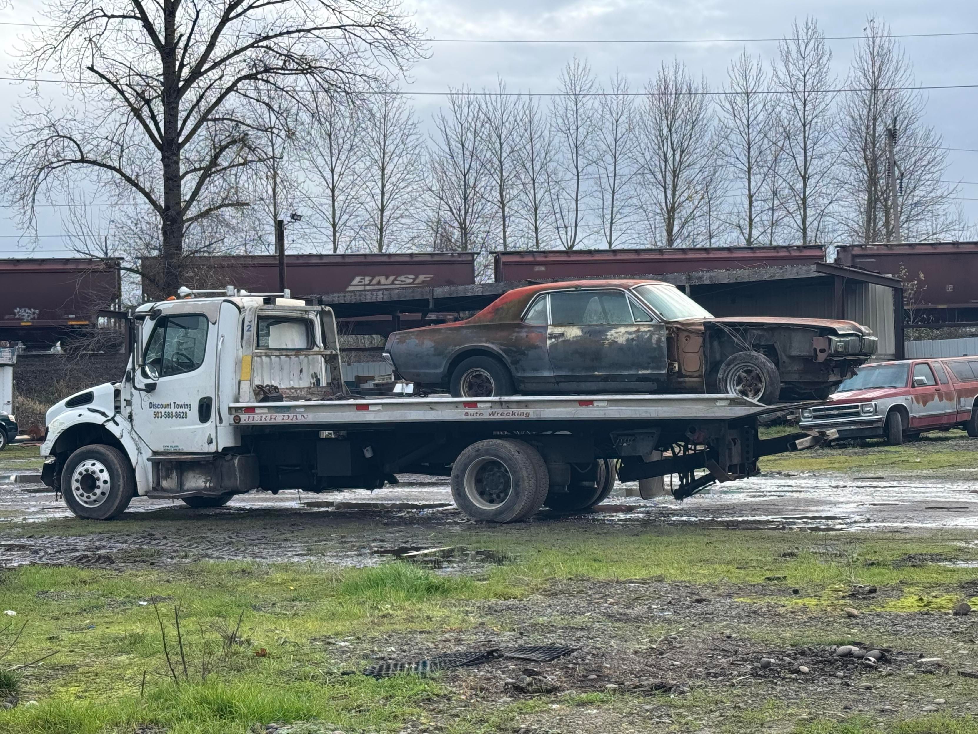 A tow truck is carrying a rusty, vintage car with a partially damaged body. In the background, there are several abandoned vehicles and a train track with freight cars. The scene is set in a muddy area with sparse vegetation.