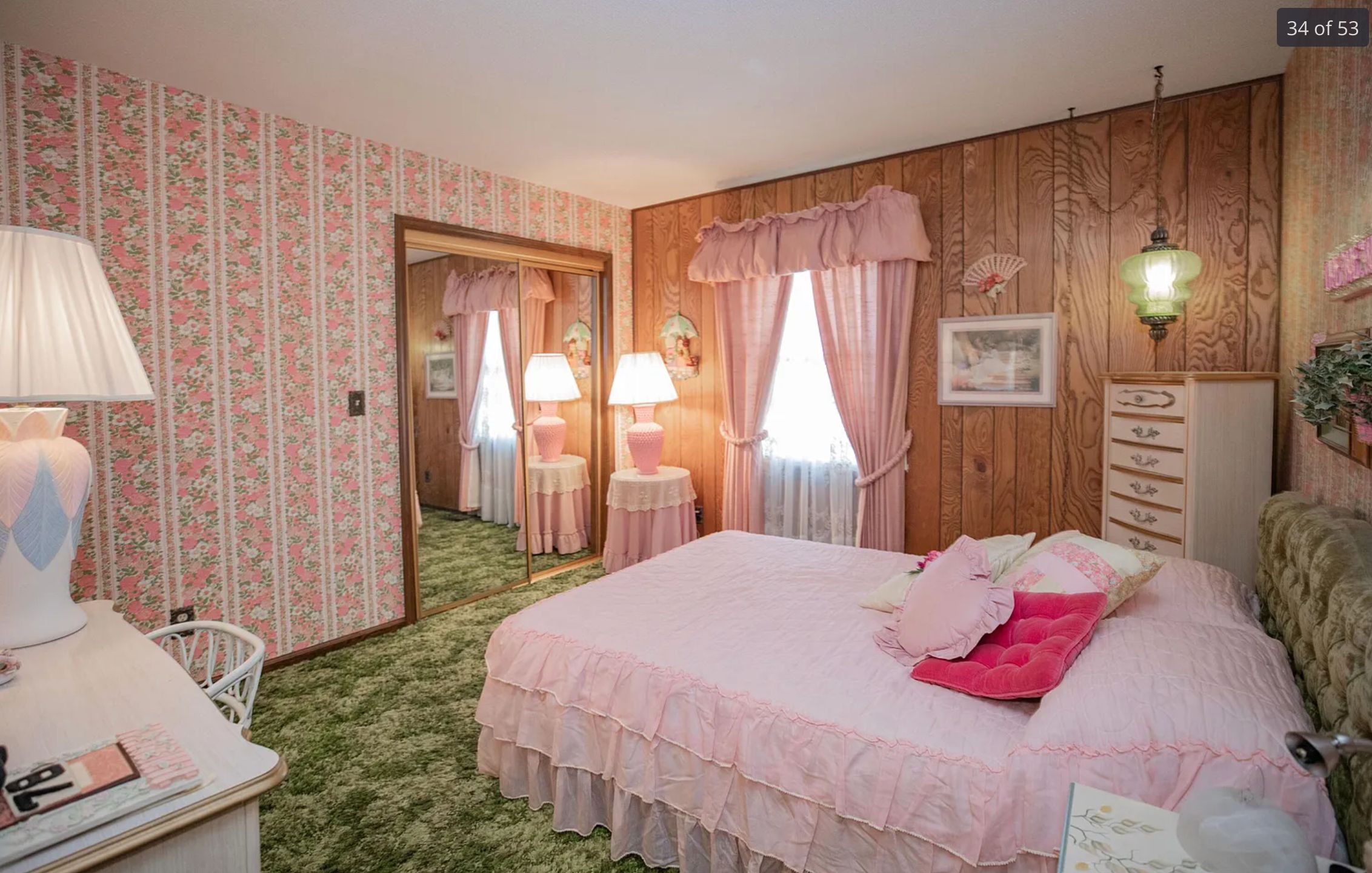 A bedroom that is also very 1970s with patterned carpet in shades of green, a wall with striped floral wallpaper, another wall with wood paneling, and a bed with a frilly pink cover