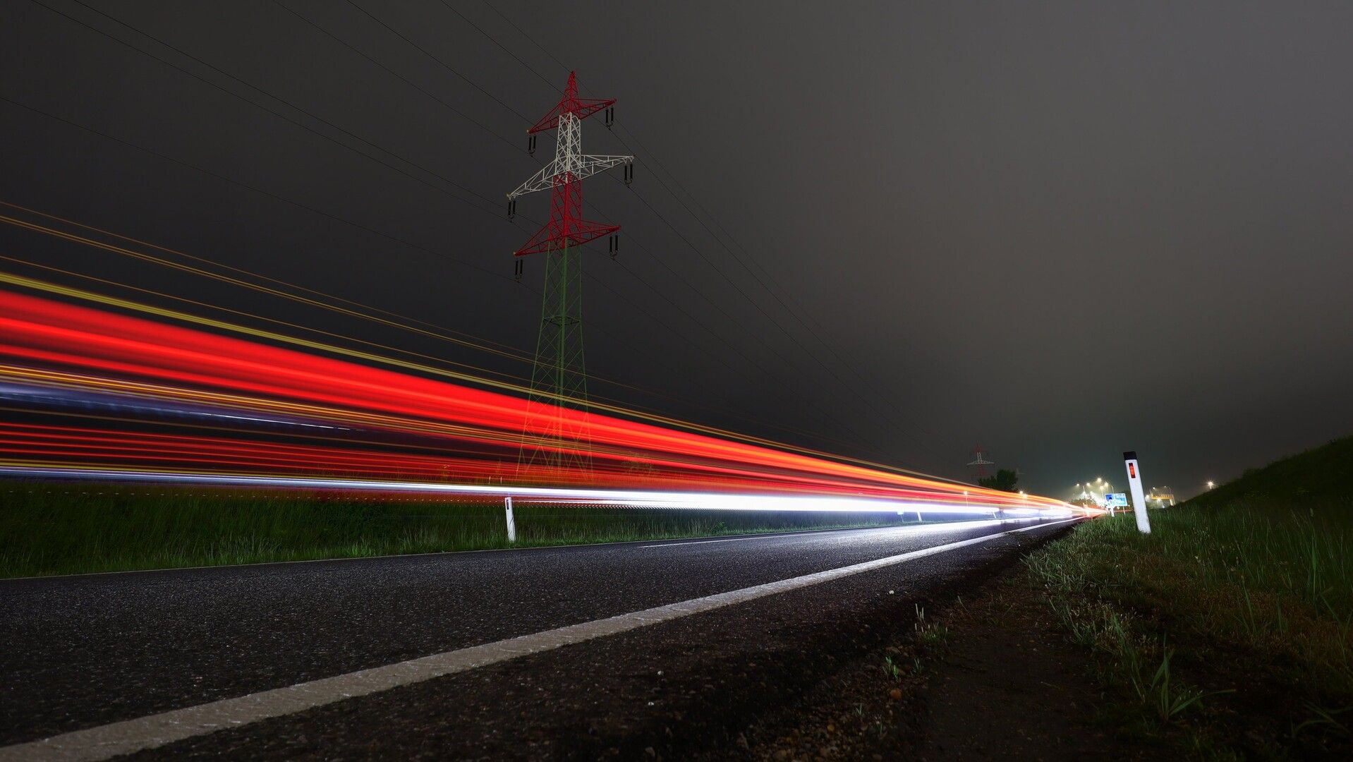 Langzeitbelichtungen auf einer dunklen und leicht vernebelten Bundesstraße. Die Scheinwerfer und Rücklichter von zahlreichen Fahrzeugen ziehen mehrere Lichtstreifen durch das Bild. Das Foto ist sehr weitwinkelig aus Bodennähe aufgenommen. Im Hintergrund sieht man einen rot-weiß-roten Hochspannungsmasten, in der Ferne Straßenbeleuchtung bei einem Kreisverkehr.