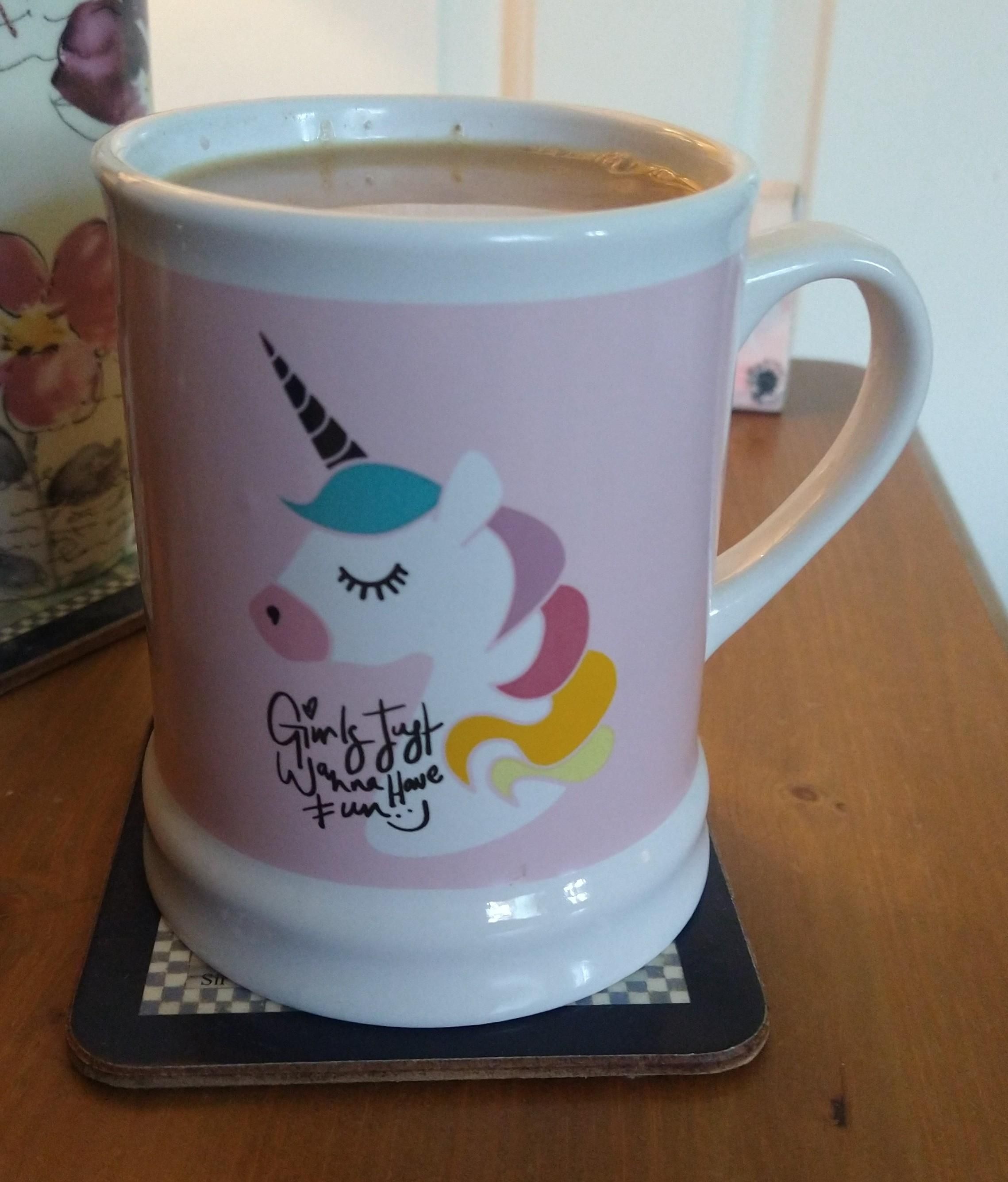 Large tankard shaped mug of tea on a bedside cabinet, it has a pink background with a unicorn head with multicoloured mane 