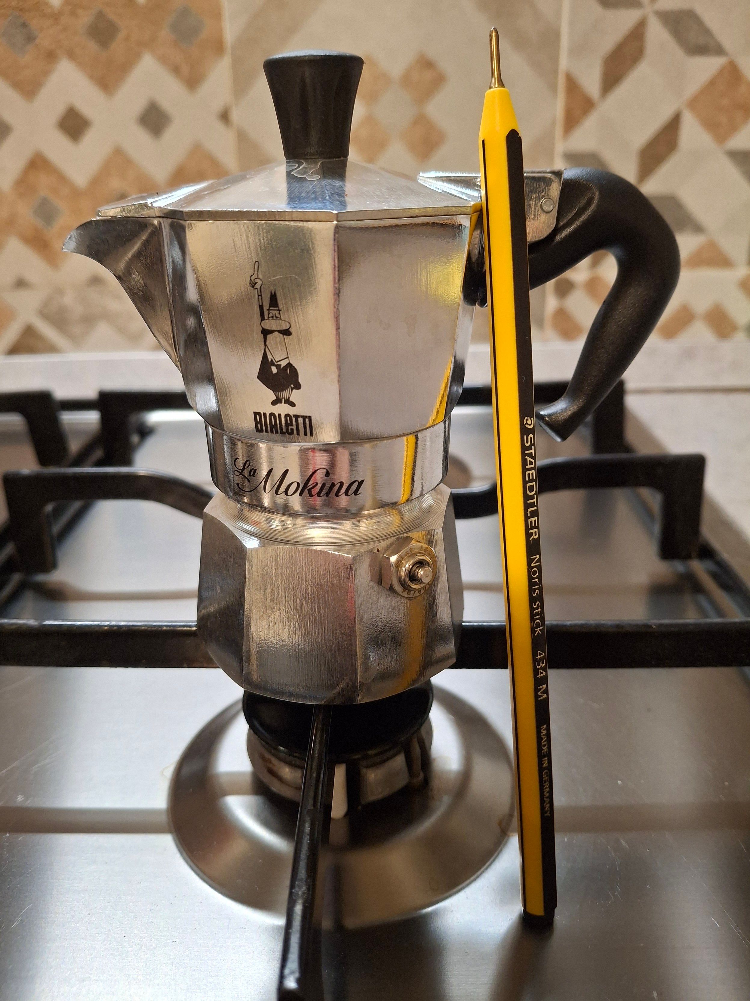 Sul fornello più piccolo di un piano cottura in acciaio a gas, una caffettiera in alluminio da mezza tazza con manico e pomello nero con impresso il logo Bialetti e la scritta in corsivo elegante "La Mokina". Accanto alla caffettiera una penna gialla e nera "STAEDTLER Noris stick 434 M MADE IN GERMANY"