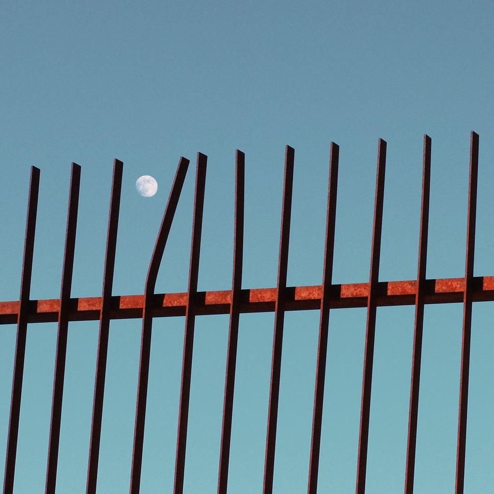 une grille dressée devant le ciel, la lune entre deux barreaux tordus
