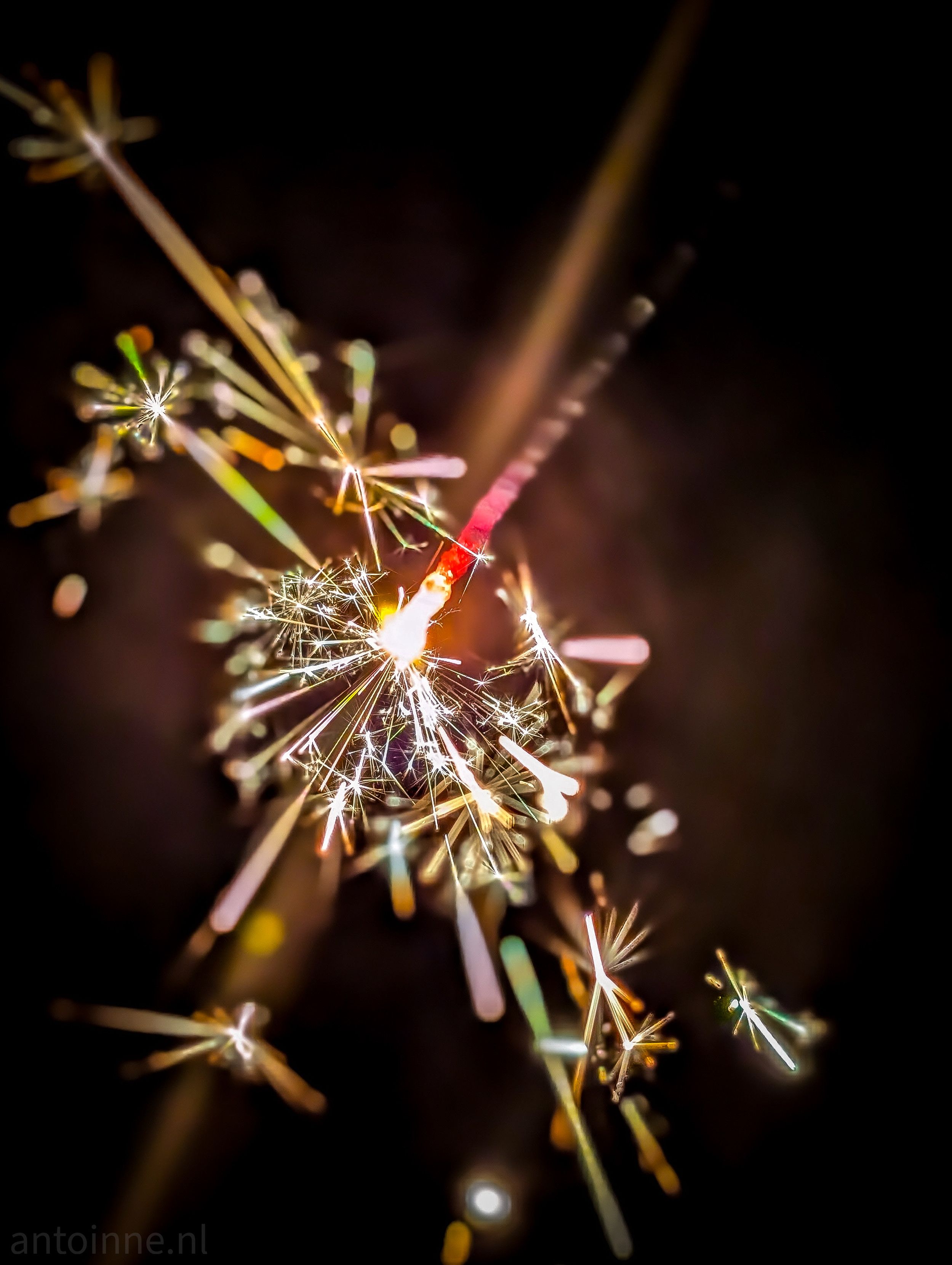 A high-contrast macro photograph of a lit sparkler against a dark, out-of-focus background. The core of the sparkler is a bright, white-hot glow where the chemical reaction is most intense. 

From this center, a red-hot metal rod extends diagonally upward toward the right. Countless sparks radiate outward in various directions. The palette is dominated by warm tones (glowing whites, oranges, and reds).