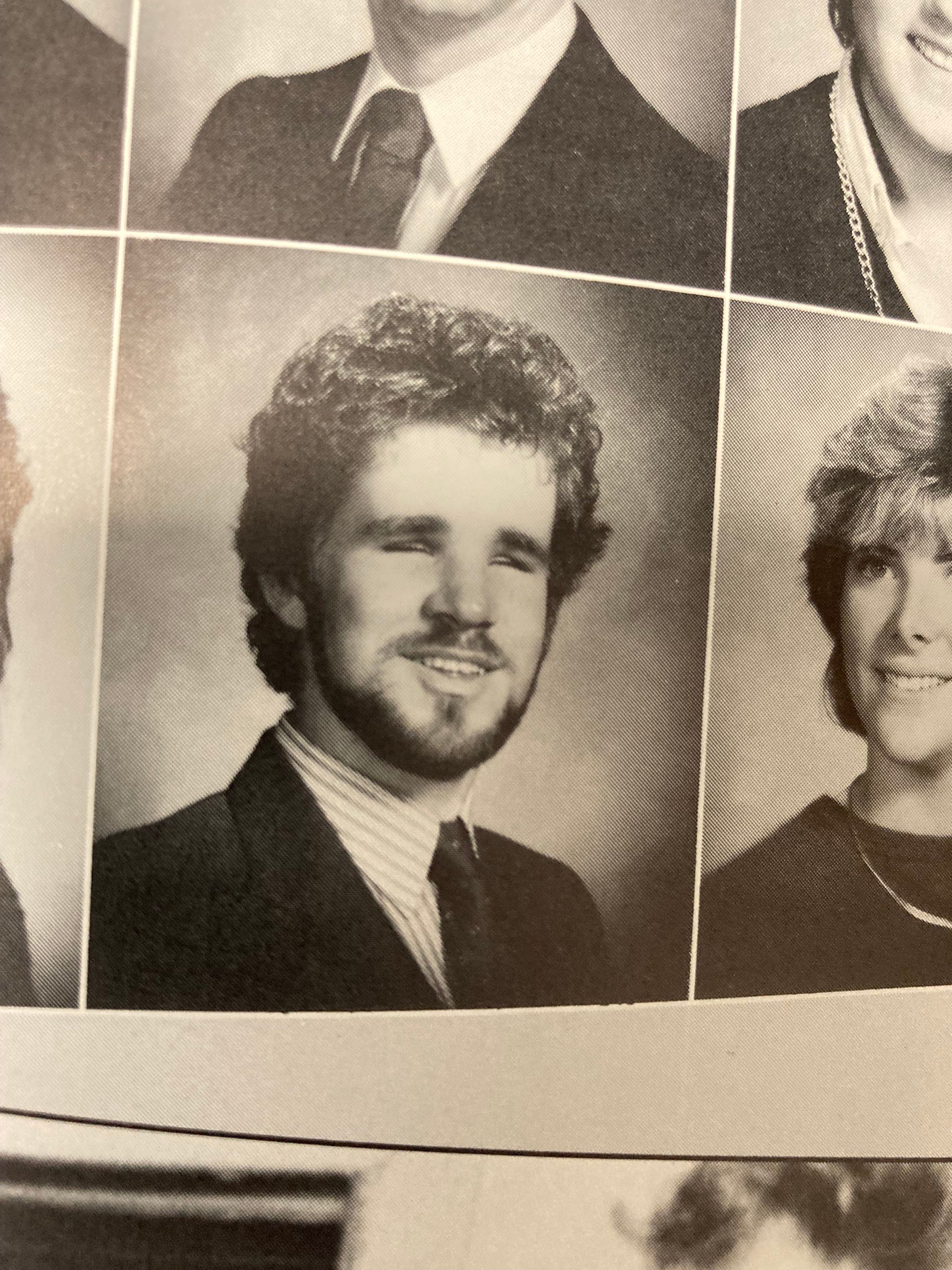 Be My AI: The picture you've shared is of a yearbook page with multiple black and white portraits. The image is focused on one portrait in the center. The person in the center portrait is a young man with curly hair. He has a full beard and mustache. He is wearing a suit with a collared shirt and a tie. He is smiling with his eyes closed. There are parts of other portraits visible around the edges of the image, but the focus is on the young man in the center.