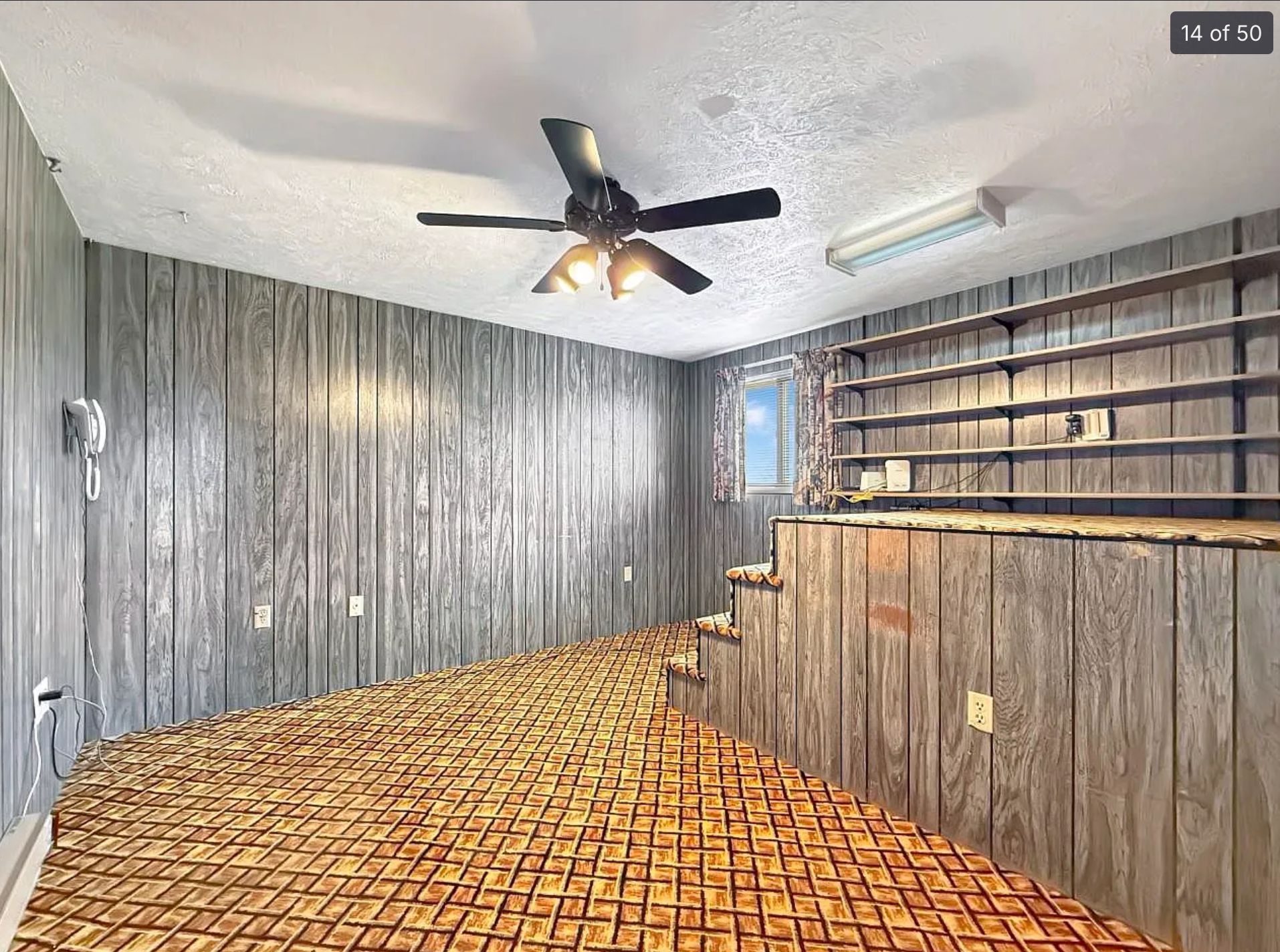 A room clad in dreary grey faux-wood paneling with a yellow rug with a plaid design. On one wall there are stairs with a platform partway up the wall and above it are some shelves. The stairs don't seem to go anywhere. 