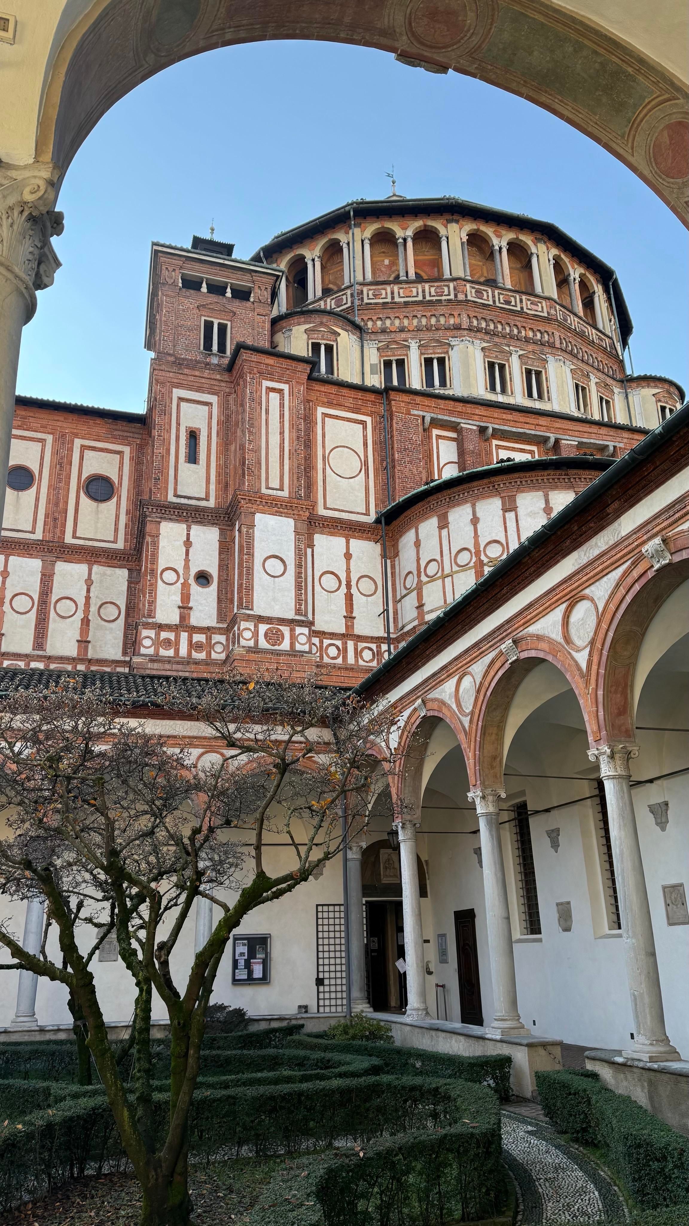 A view of the beautiful Church and Dominican Convent of Santa Maria delle Grazie in Milan. It is an historic building featuring intricate brickwork, circular patterns, and ornate arches. The image captures a courtyard with neatly trimmed hedges and a tree, framed by classical columns. The sky is clear and blue, highlighting the architectural details.
