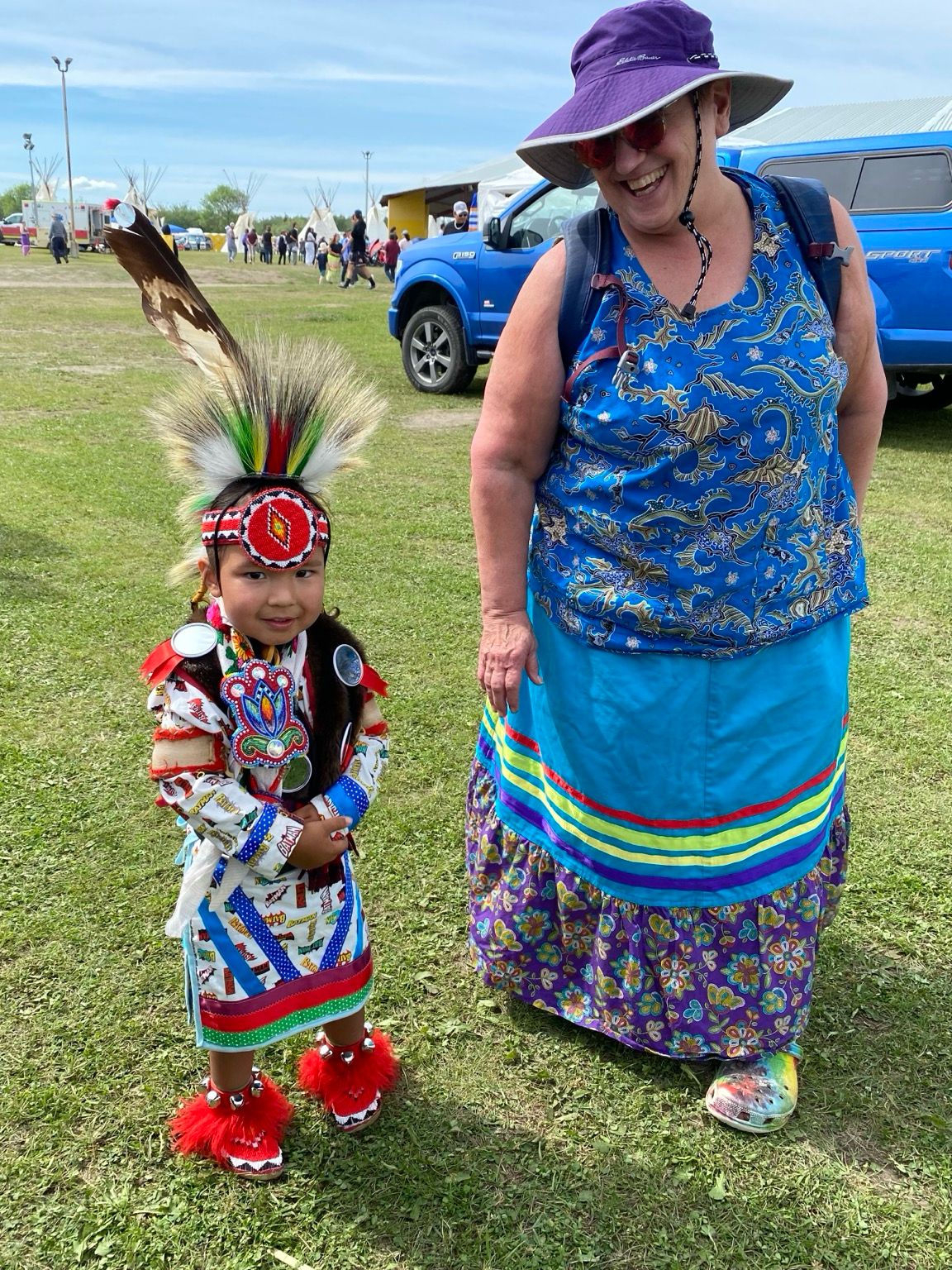 A woman wearing a turquoise ribbon skirt stands next to a tiny pow wow dancer in Batman regalia 