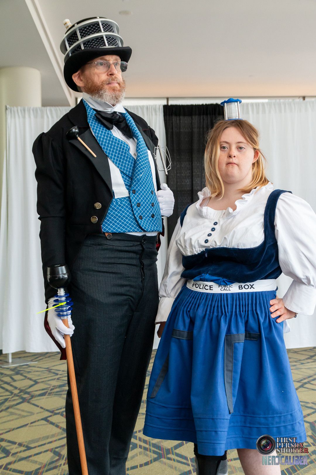 A man cosplaying as a Dapper Dalek wearing a full suit, tailcoat and top hat next to a young woman with Down syndrome cosplaying as a TARDIS