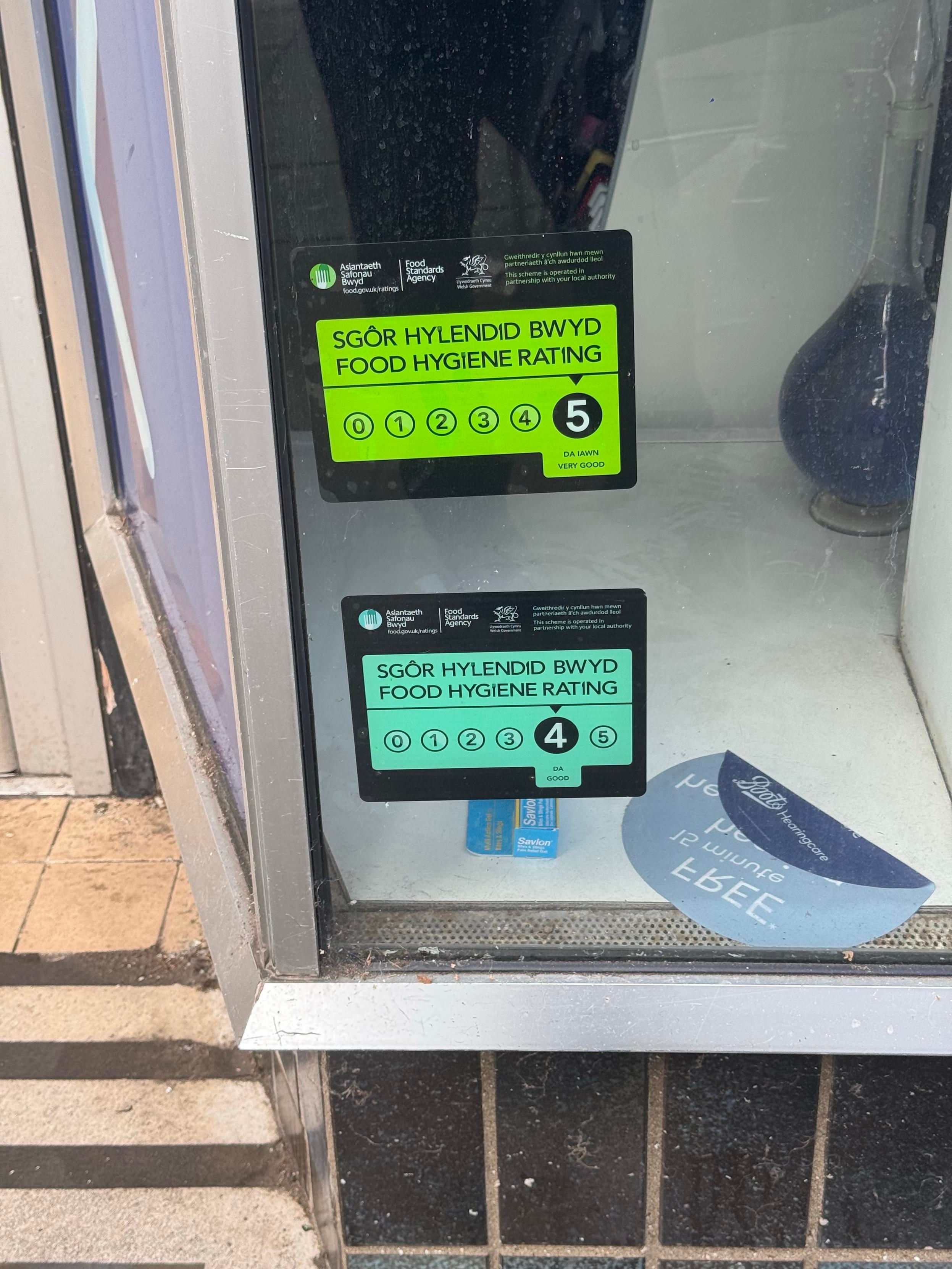 A window displaying two food hygiene rating stickers. The top sticker shows a rating of 5 (very good), while the bottom sticker shows a rating of 4 (good). Both are in Welsh and English.