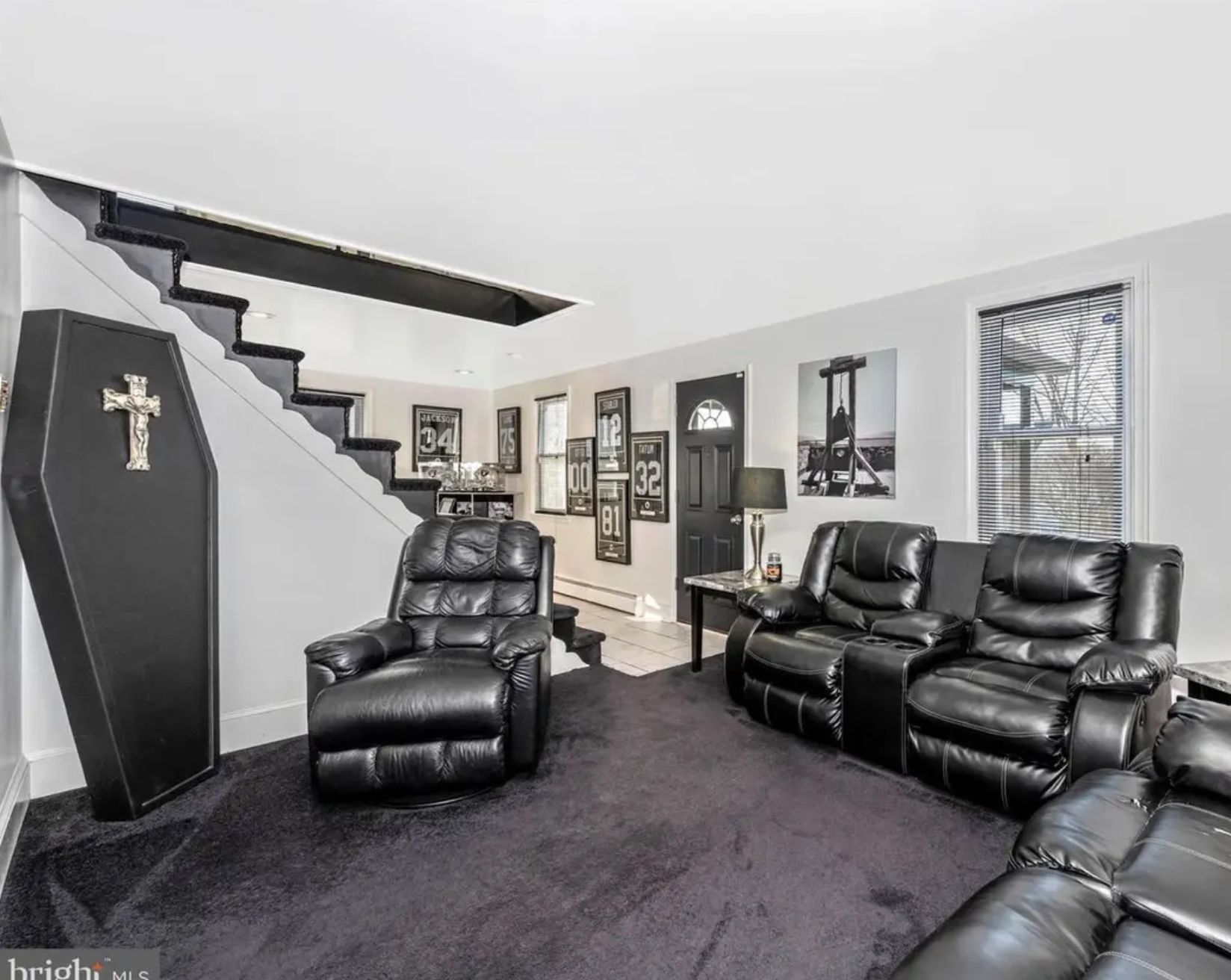 A living room with a black carpet and black leather chairs. In the corner is a black fake casket with a cross on it