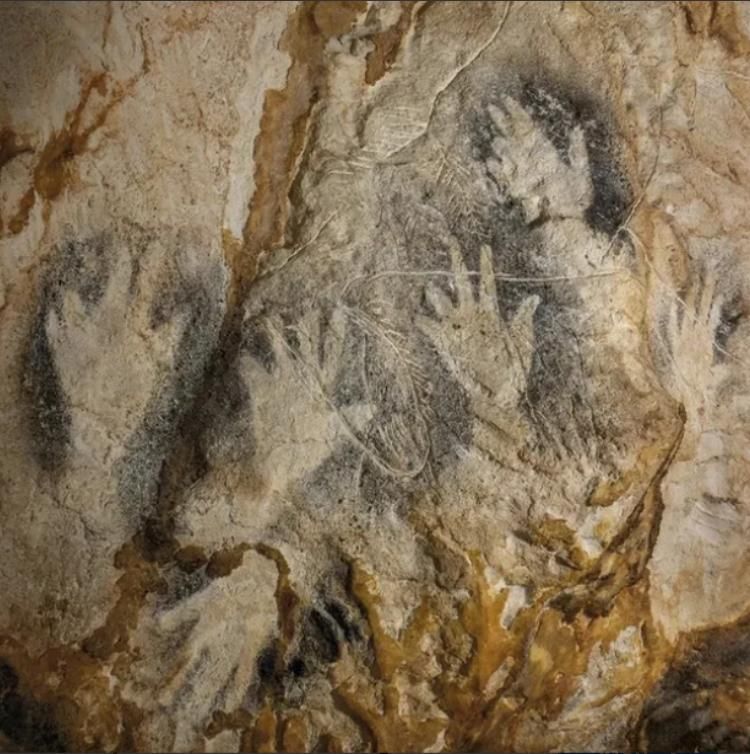 Cave painting of several hands. The paintings are made by placing your hand on the cave wall and blowing pigment into them with a straw. The pigment in this case is black. Many of these hand prints have shortened or missing fingerprints; there are a few different interpretations, some very silly (ritual amputation) but some like the top left one are almost unmistakably made by someone making a different shape with their hand, not someone missing fingers 