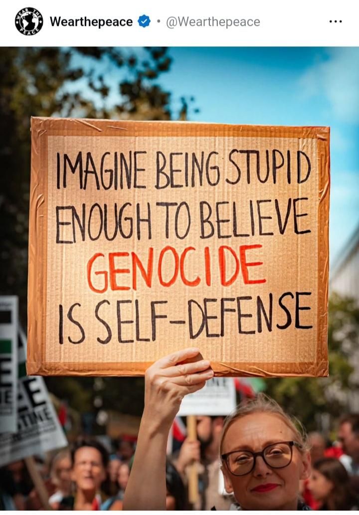 A woman wearing glasses holds a sign that says: " Imagine being stupid enough to believe GENOCIDE is self defense"