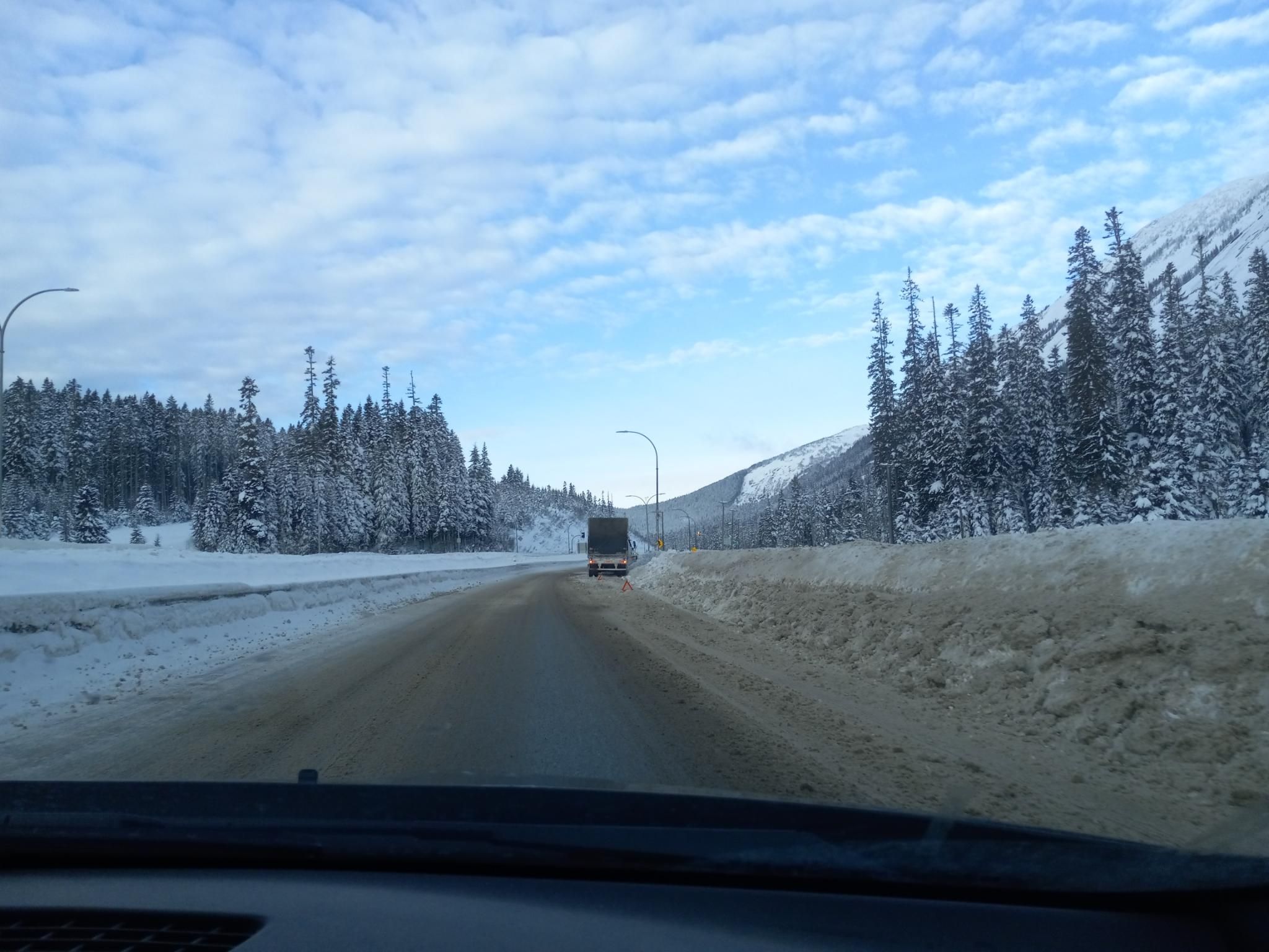 Looking along a muddy, slushy highway road with lots of snow on the sides toward a somewhat smashed semi-truck on the shoulder up ahead.  There are tall pines on both sides of the road with snow, and tall mountains.  The sky above is blue with some clouds