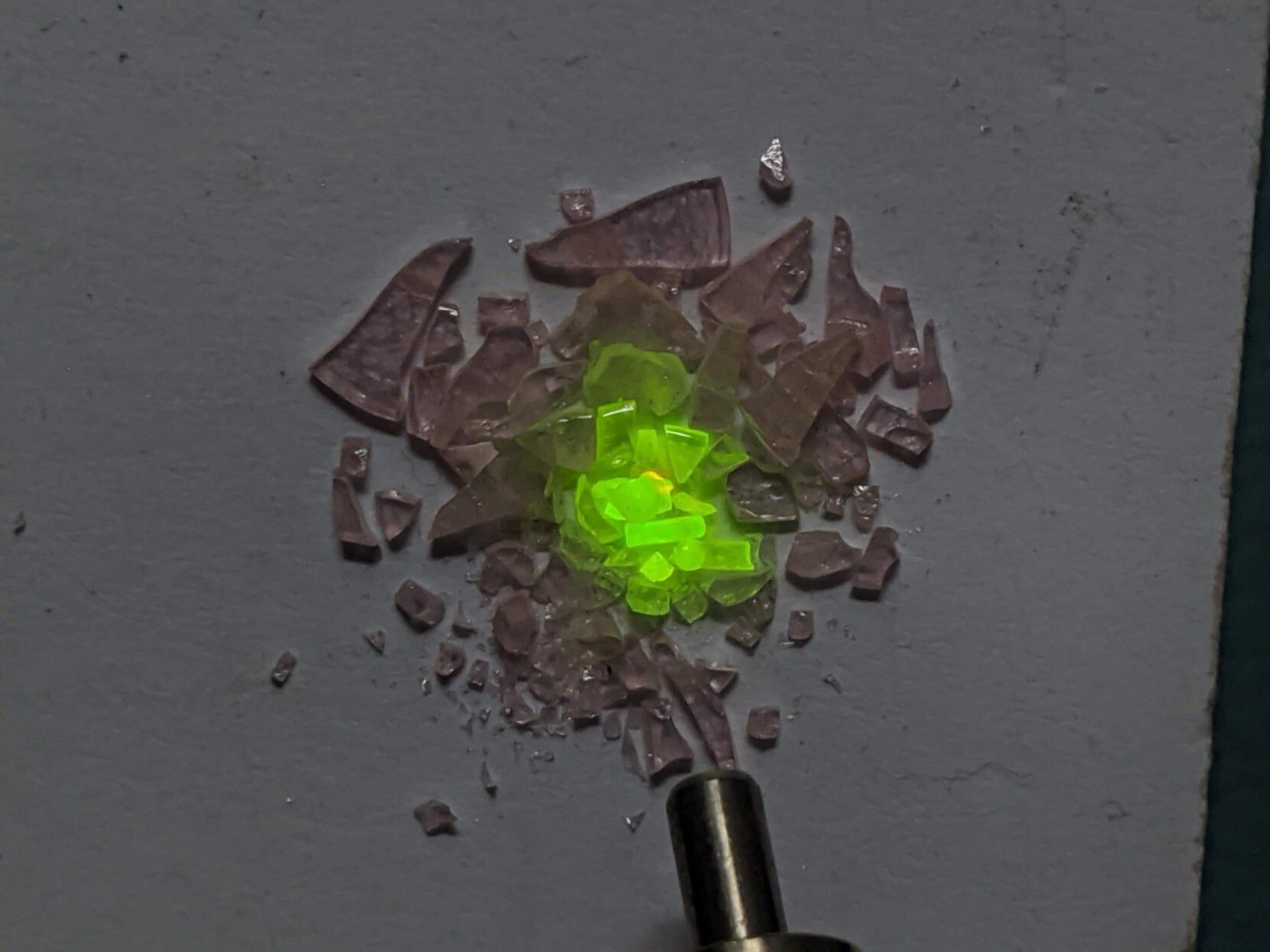 A heap of pink glass fragments. The tip of an optical fibre outputting infrared light is visible in the lower half of the image. Where the infrared light hits the glass fragments they fluoresce a bright green.