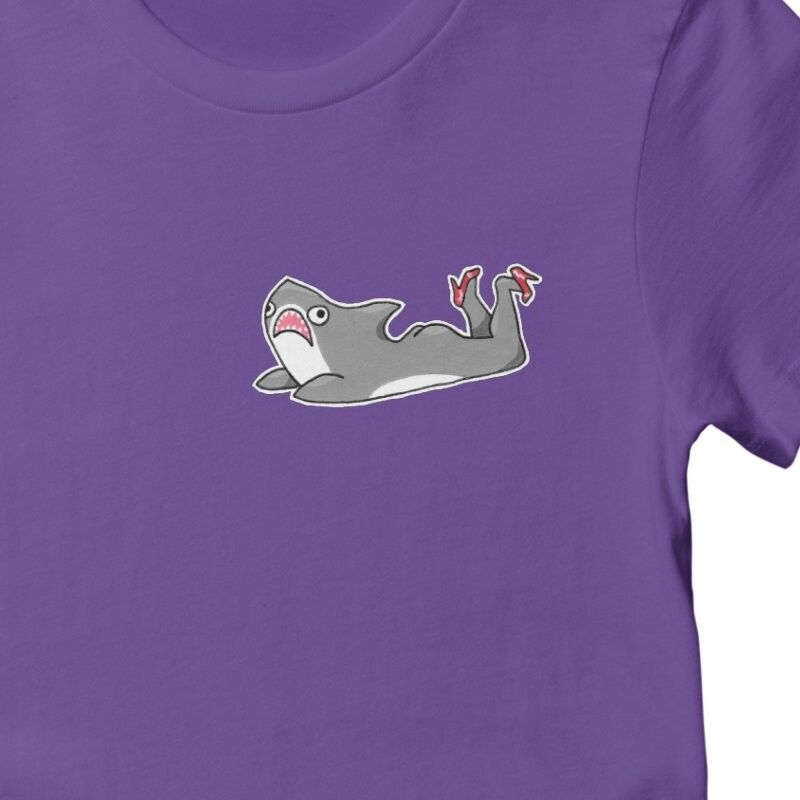 A closeup of the top left corner of a women's purple t-shirt with a small image of a grey cartoon shark with legs, wearing red high heels. It's the silly doodle I drew the other day, made manifest