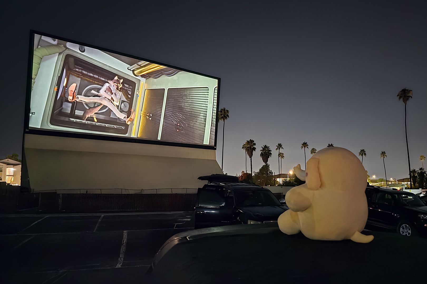 Photo of the Creature Mastodon stuffed toy sitting on a car at a drive-in theater during a late-night showing of The Bad Guys 2. The Creature, towards image-right, is sitting on a black moving blanket atop a black car, facing away from the camera as it faces a drive-in theater projector screen. On the projector screen in the background, towards image-left, is Mr. Wolf with his legs spread and arms crossed as he floats in the air, a still from a showing of The Bad Guys 2. In the midground, all around the Creature, are other moviegoers' cars with their trunks pointed toward the projector screen as well.