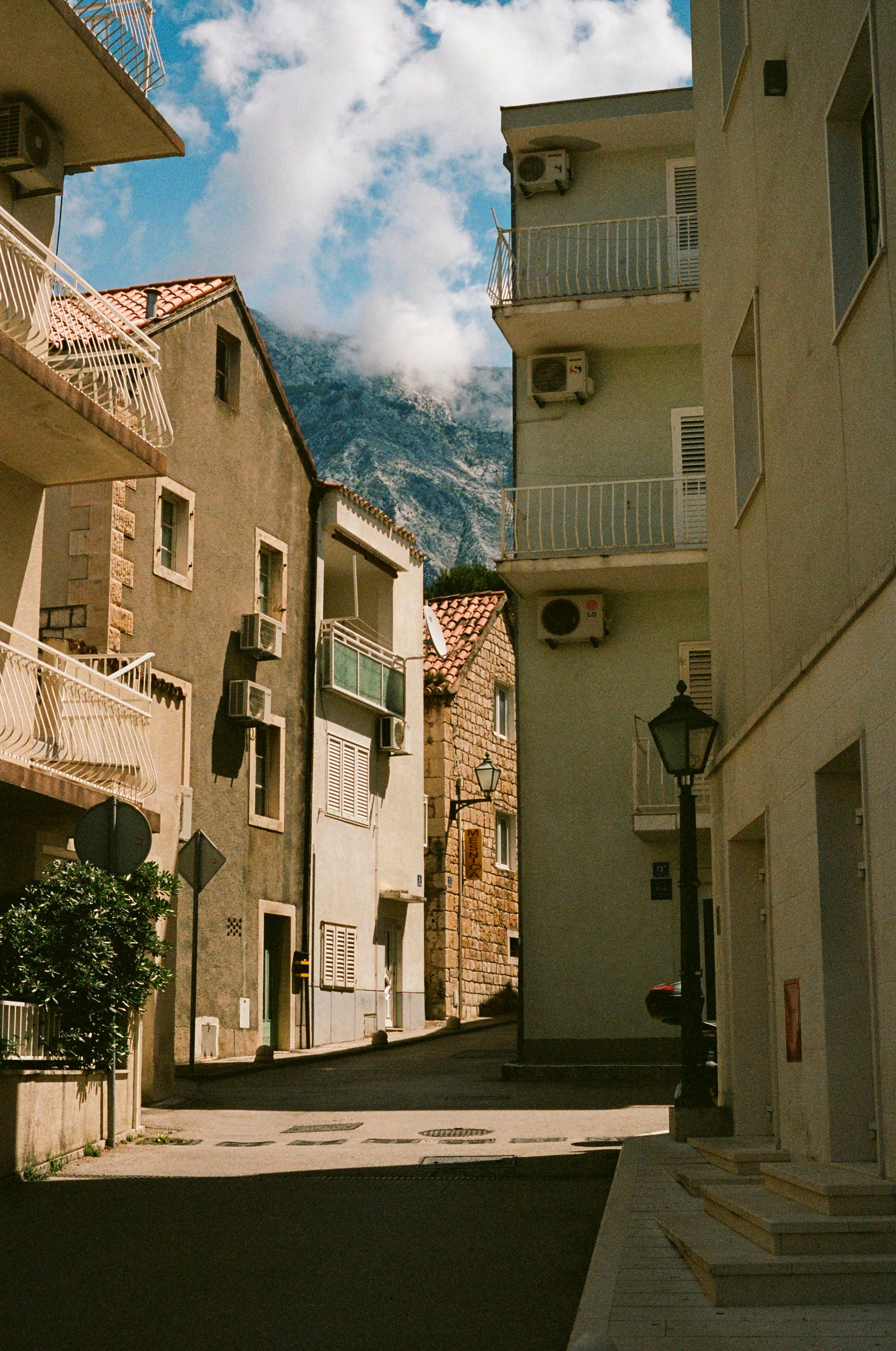 A street of Mediterranian houses, half in shadow, half in sunlight, with a tall mountain peaking out behind it, confidently grazing passing by clouds.