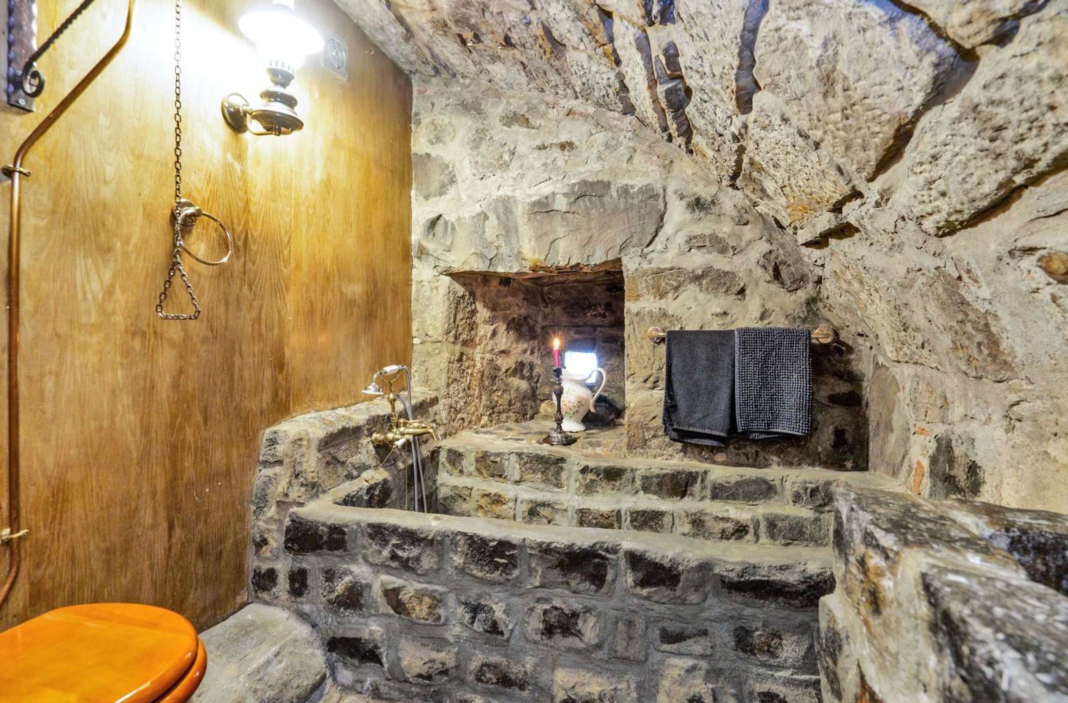 A bath made of stone blocks in a room with a curved stone ceiling and the back wall is made of wood. The bath does not look comfortable. 