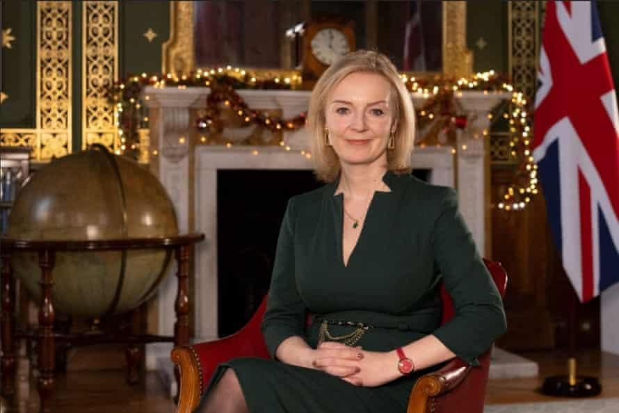 Yes she was prime minister! Liz Truss. Here we see her as Roweley Birkin QC from the Fast Show. She's sat in what appears to be an exclusive racist gentleman's club. It definitely smells of cigars and cum. I'm not saying she does. She smells like salad and regret. She's wearing bank-wear and is sat in a highback leather seat with a large mirror and traditional clock on a fireplace behind her with Christmas lights and fancy gold detailing and a marble fireplace bit at the front. To one side is one of those comically large flags that look like they're designed for visually impaired people (IT'S A UNION JACK) and on the other side, one of those globe drinks cabinets that's a little too on the nose - English person with enormous privilege stashing booze inside a facsimile of the world. It's mine and I'll use it however I please. 