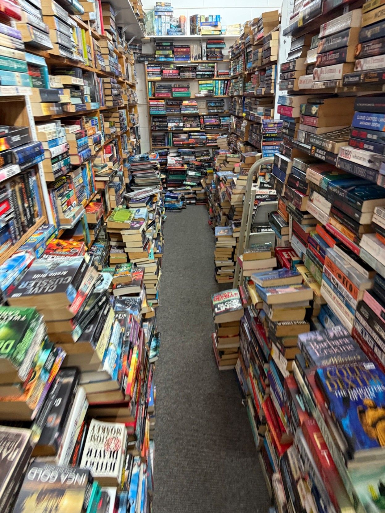 A book store with full shelves and stacked high right down to the floor on both sides of the aisle 