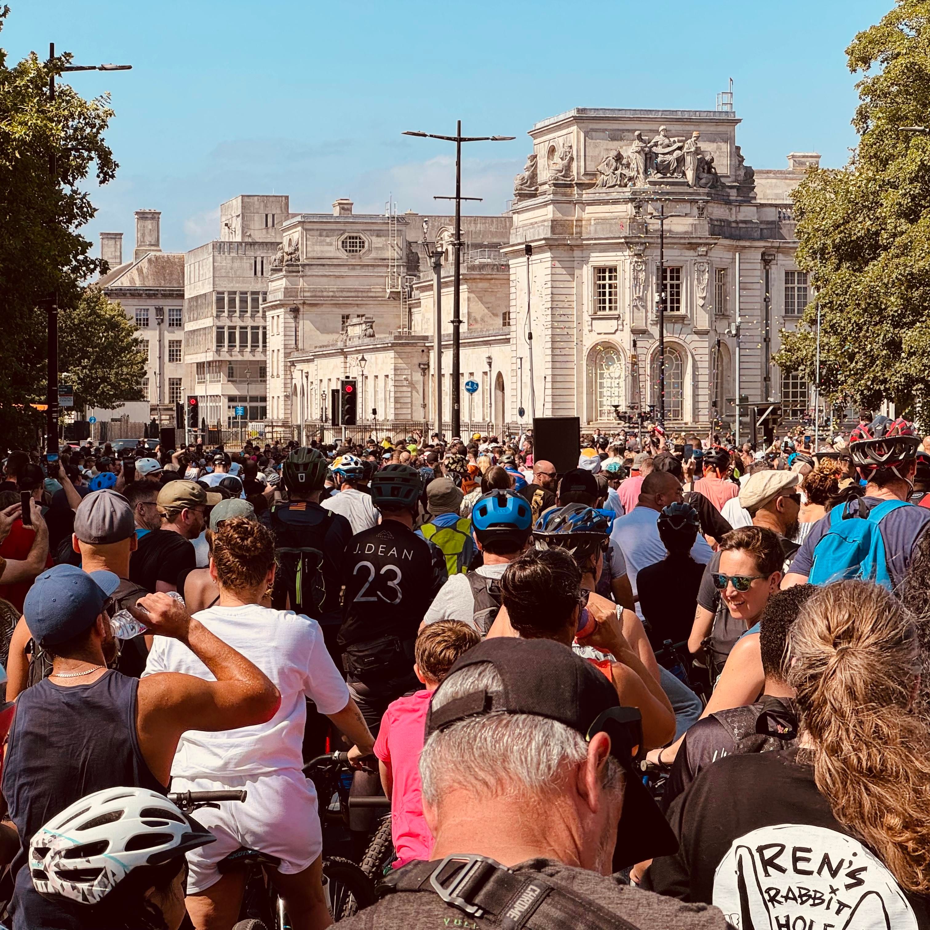 A photo depicting a crowd of hundreds of cyclists in Cardiff, attending a drum and bass group ride. In the distance, beautifully architected old buildings bask in glorious sunlight.