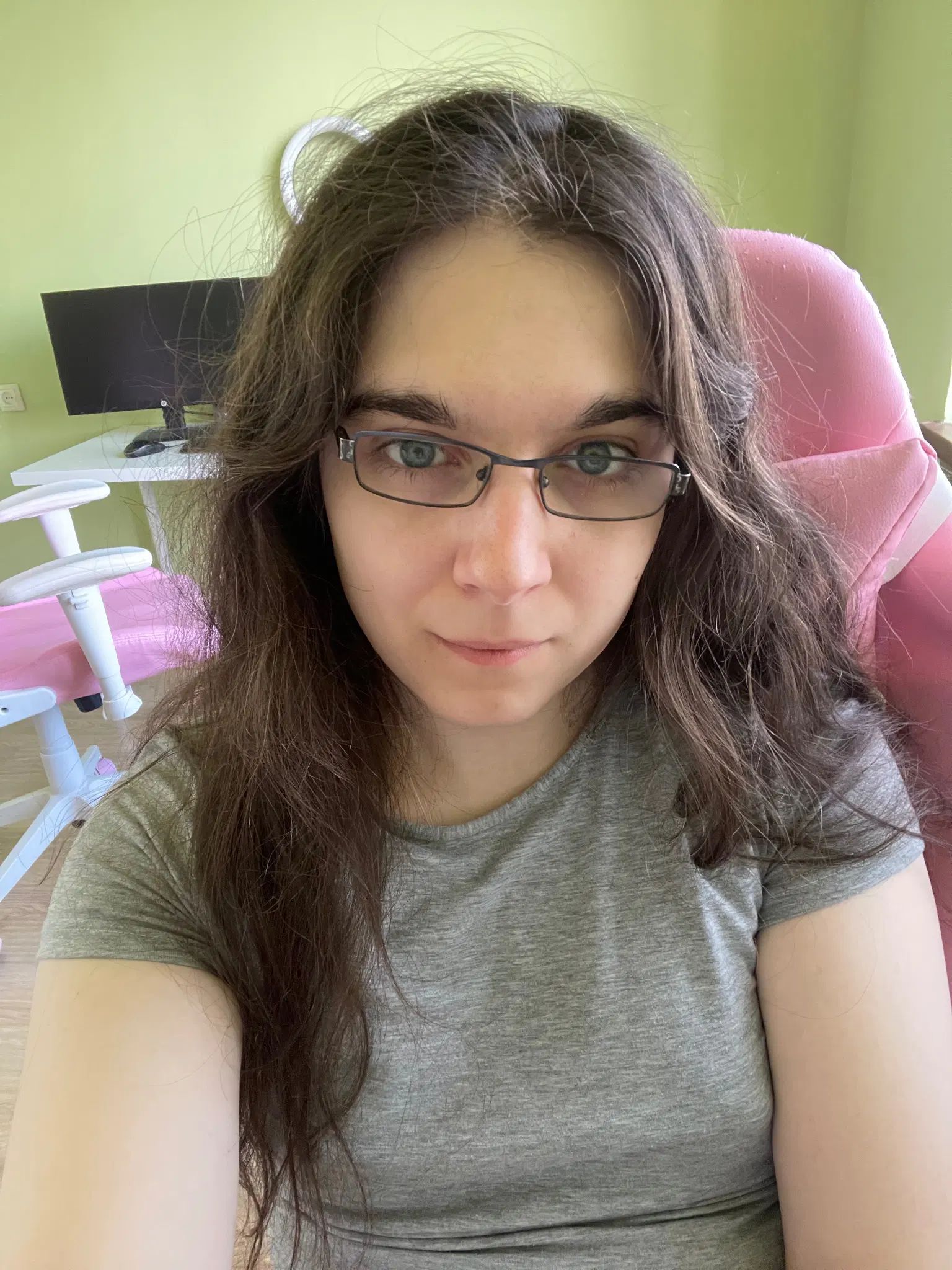 Photo of me facing forward. I'm wearing a melange grey t-shirt, rectangular glasses that are slightly crooked, and I'm sitting in a pink gaming chair. There's a second chair and a computer that's off behind me